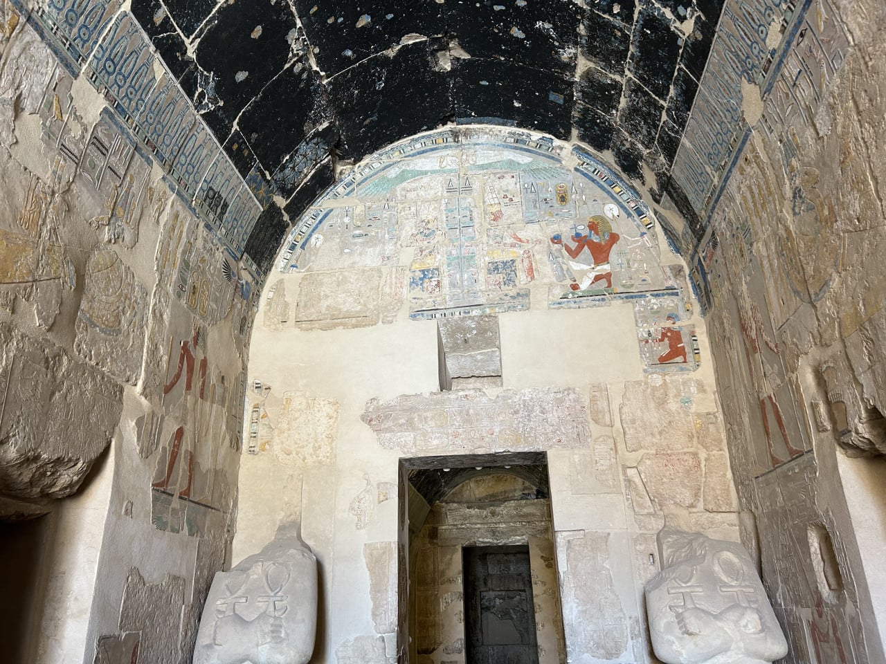 Malovaná egyptská hrobka s klenutým stropem, hieroglyfy a kamennými sochami u vchodu
