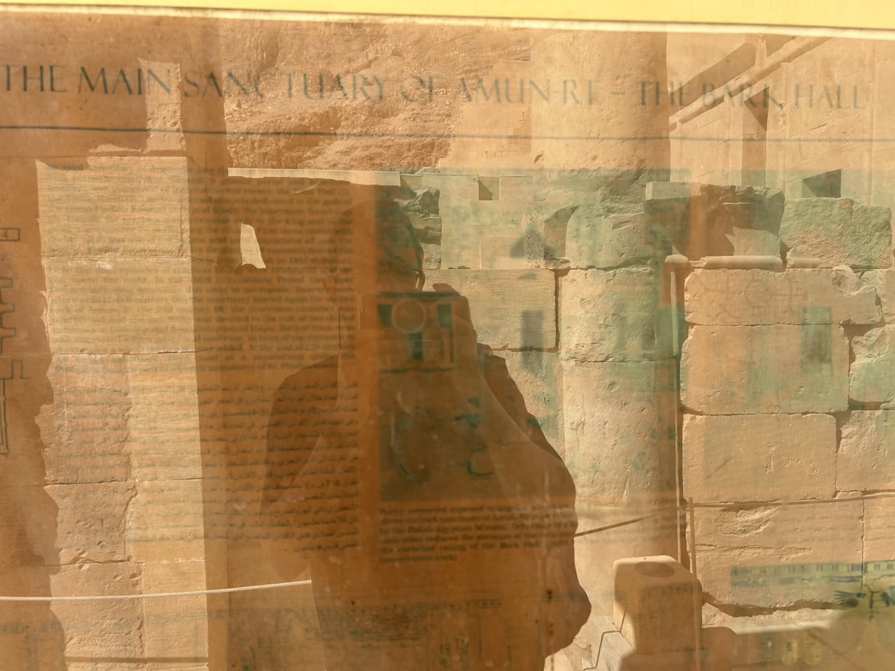 Odraz fotografa v skleněné vitríně před egyptskými kamennými zdmi a textem.