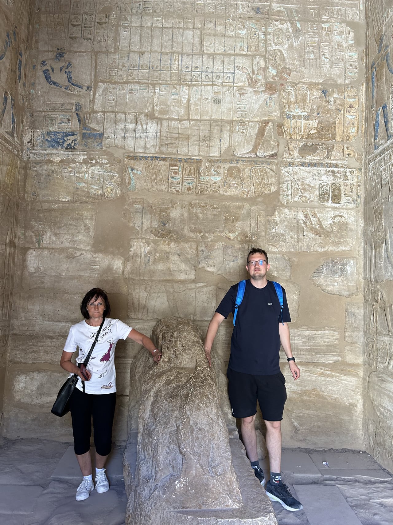Dva turisté stojí u kamenného bloku v egyptském chrámu s hieroglyfy.