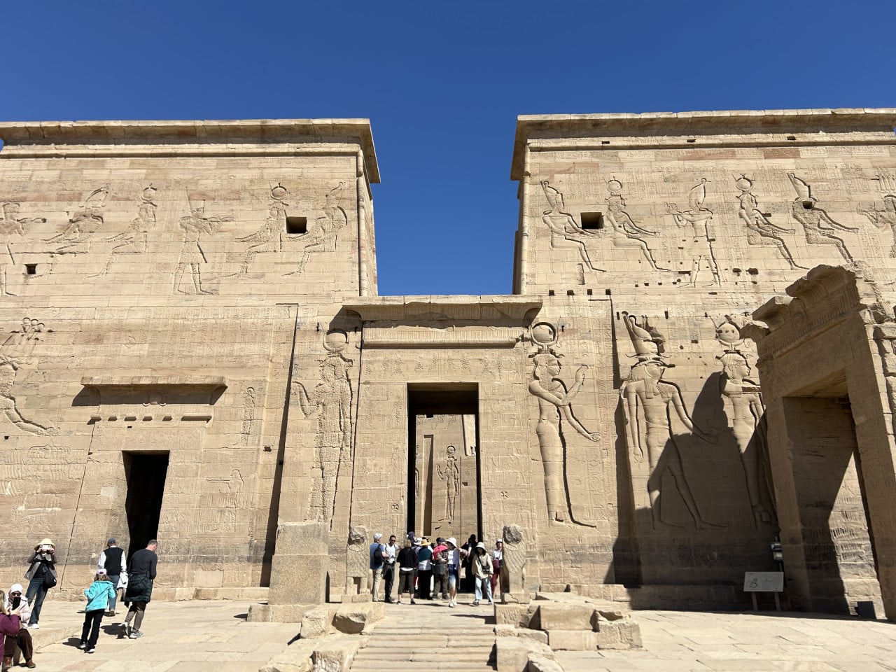 Chrámový vstup s reliéfy bohyně Isis, turisté před pískovcovou bránou pod modrou oblohou