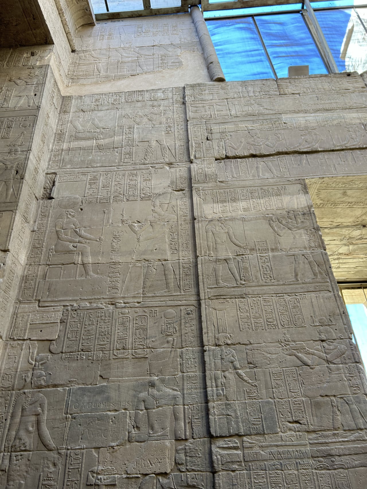 Reliéfní egyptské hieroglyfy a postavy na kamenné stěně pod modrou střešní krytinou