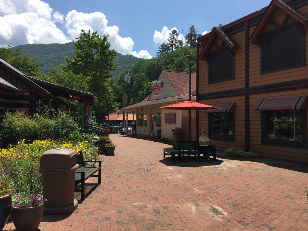 Cihlová pěší zóna s obchody, lavičkami a nápisem Village Merchant, hory v pozadí.