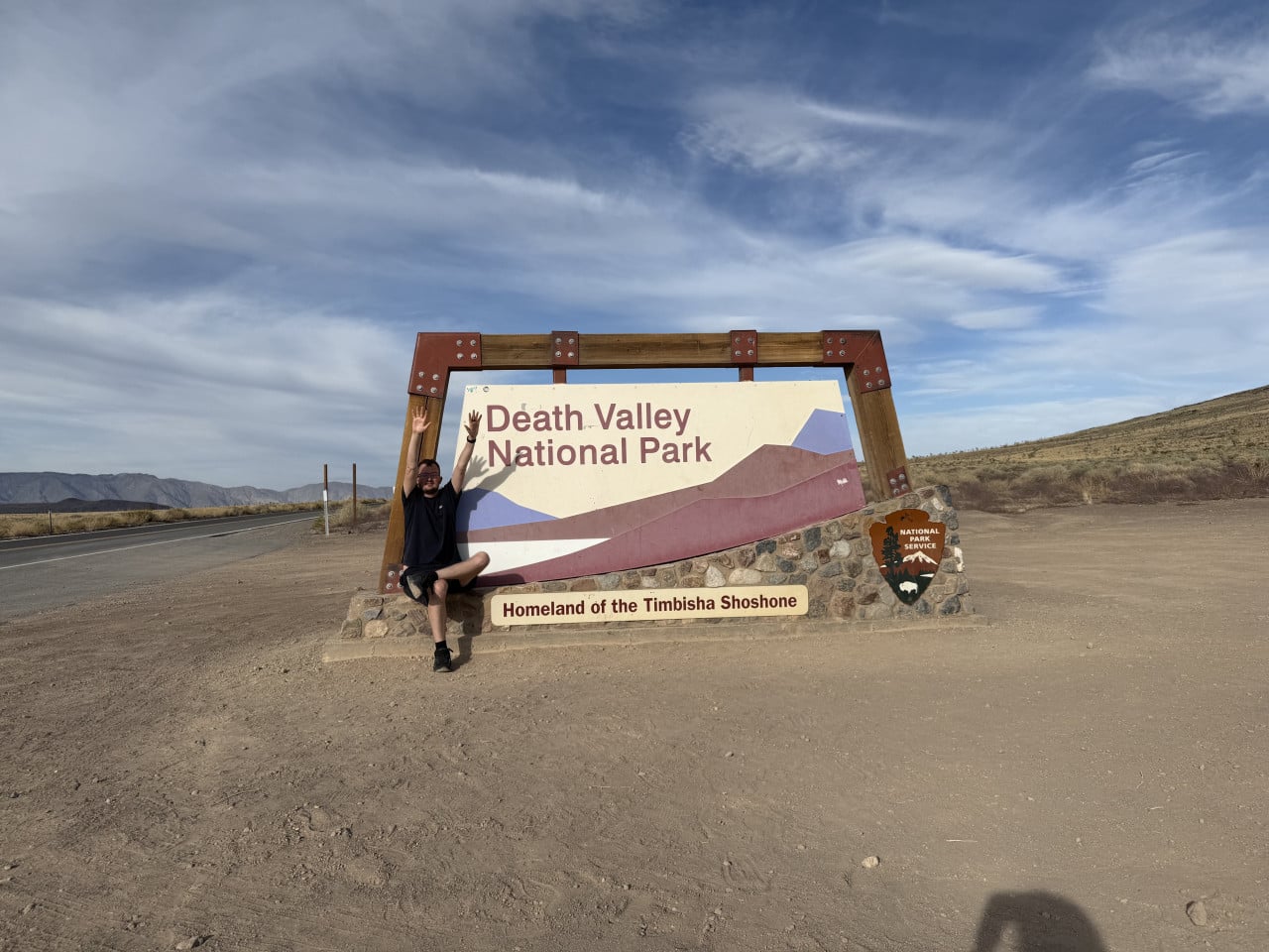 Muž sedí u cedule Death Valley National Park, ruce nahoře, pouštní krajina za ním.