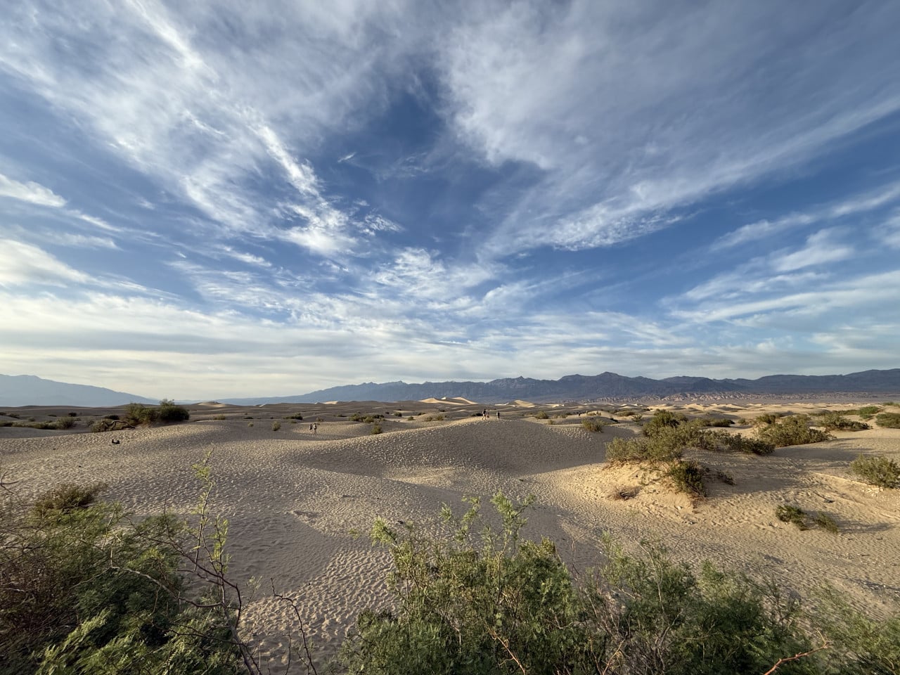 Pouštní duny v Death Valley pod dramatickou oblohou, v dálce hory a turisté.