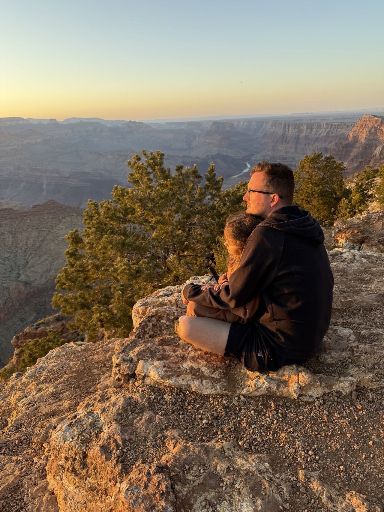 Muž s dcerou sedí na skalním okraji Grand Canyonu při západu slunce