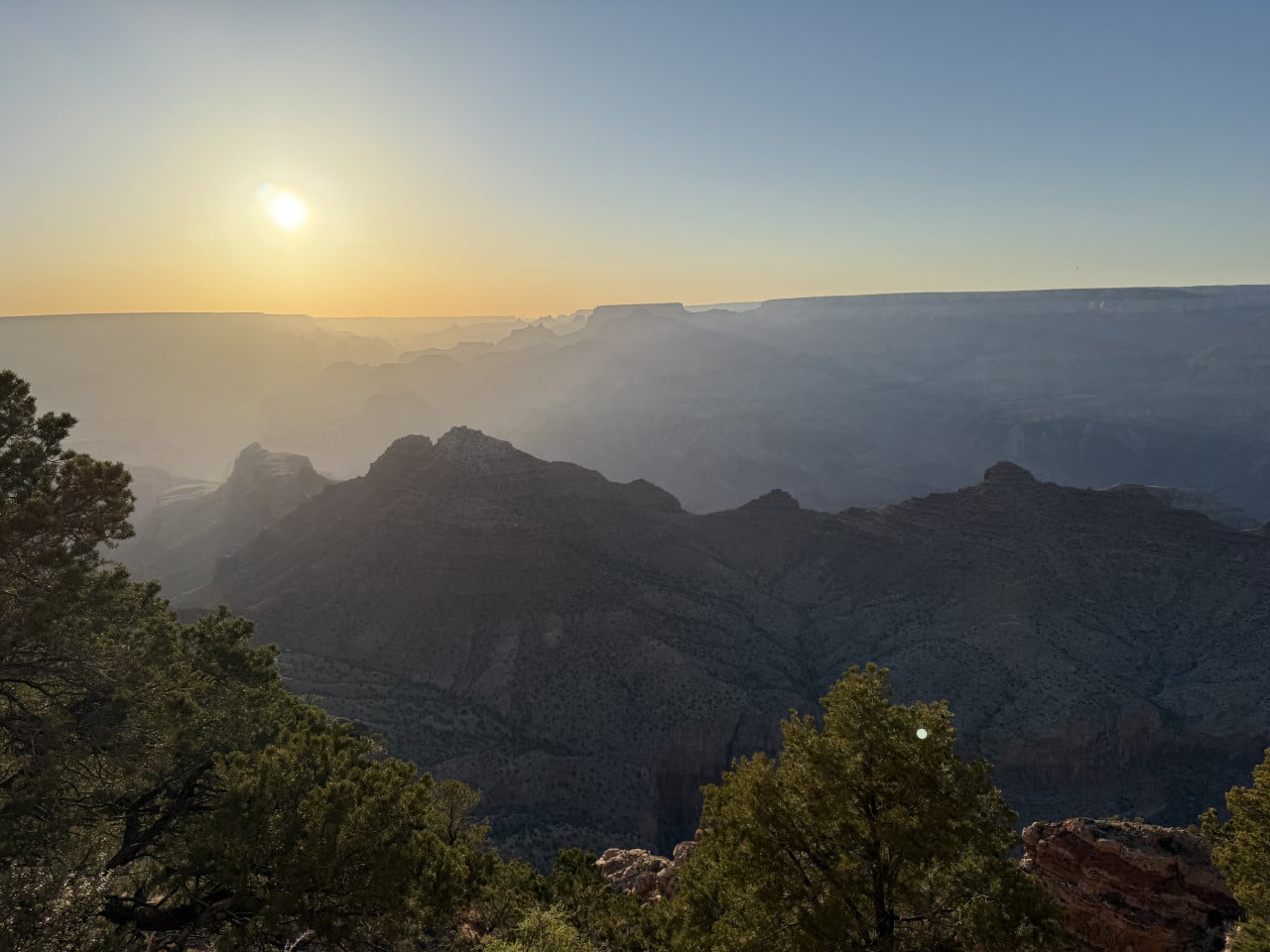 Západ slunce nad Grand Canyonem s mlhavými skalními hřebeny a stromy v popředí