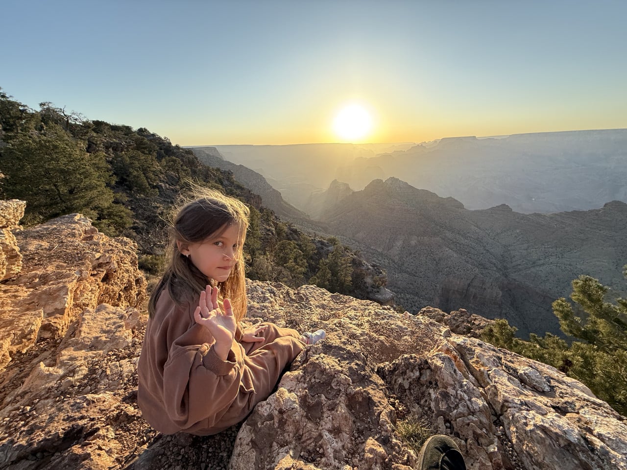 Dívka sedí na skalní římse u Grand Canyonu při západu slunce, mává do kamery.