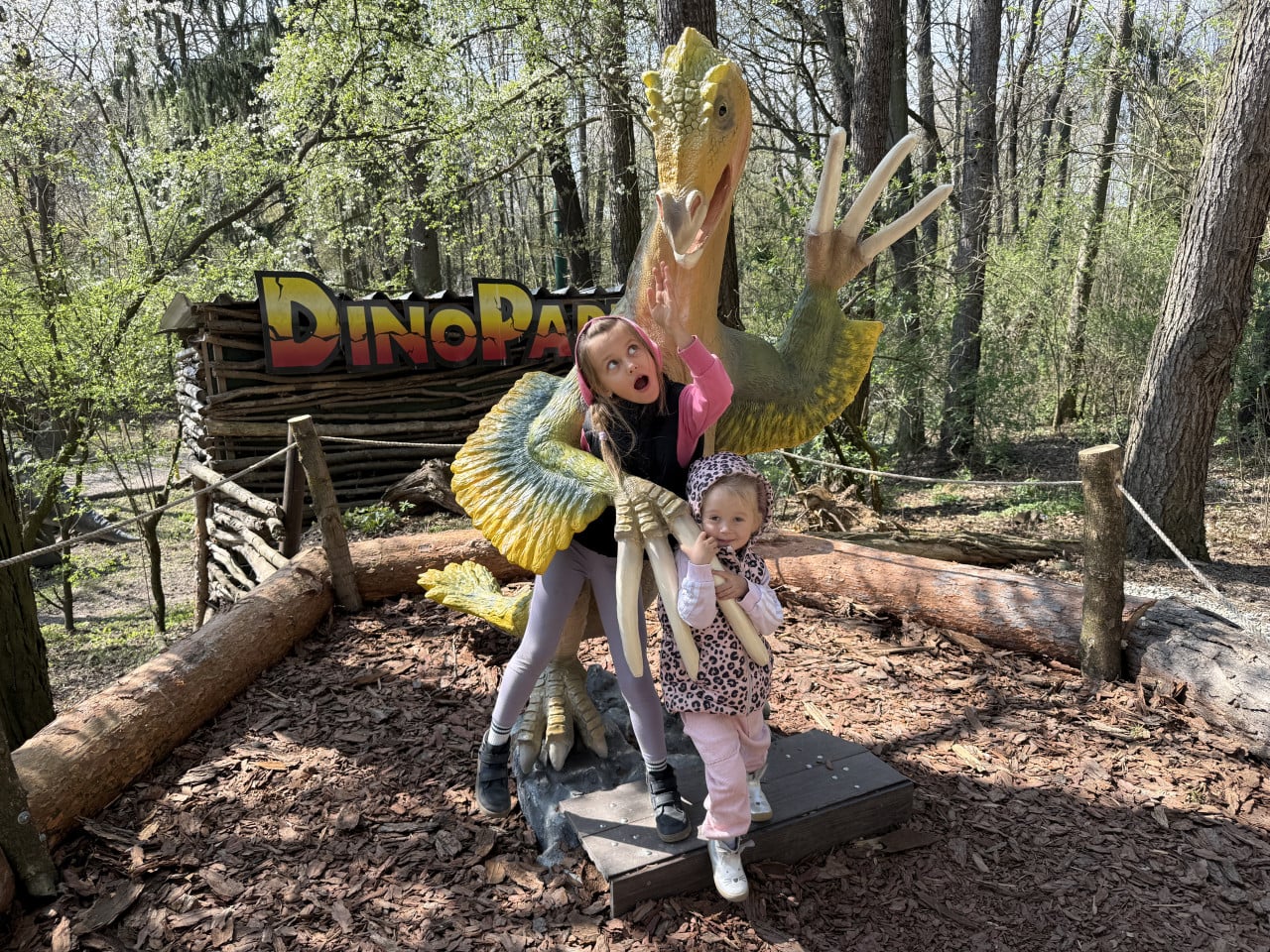 Dvě děti pózují u barevné dinosauří sochy s nápisem DinoPark v lese.