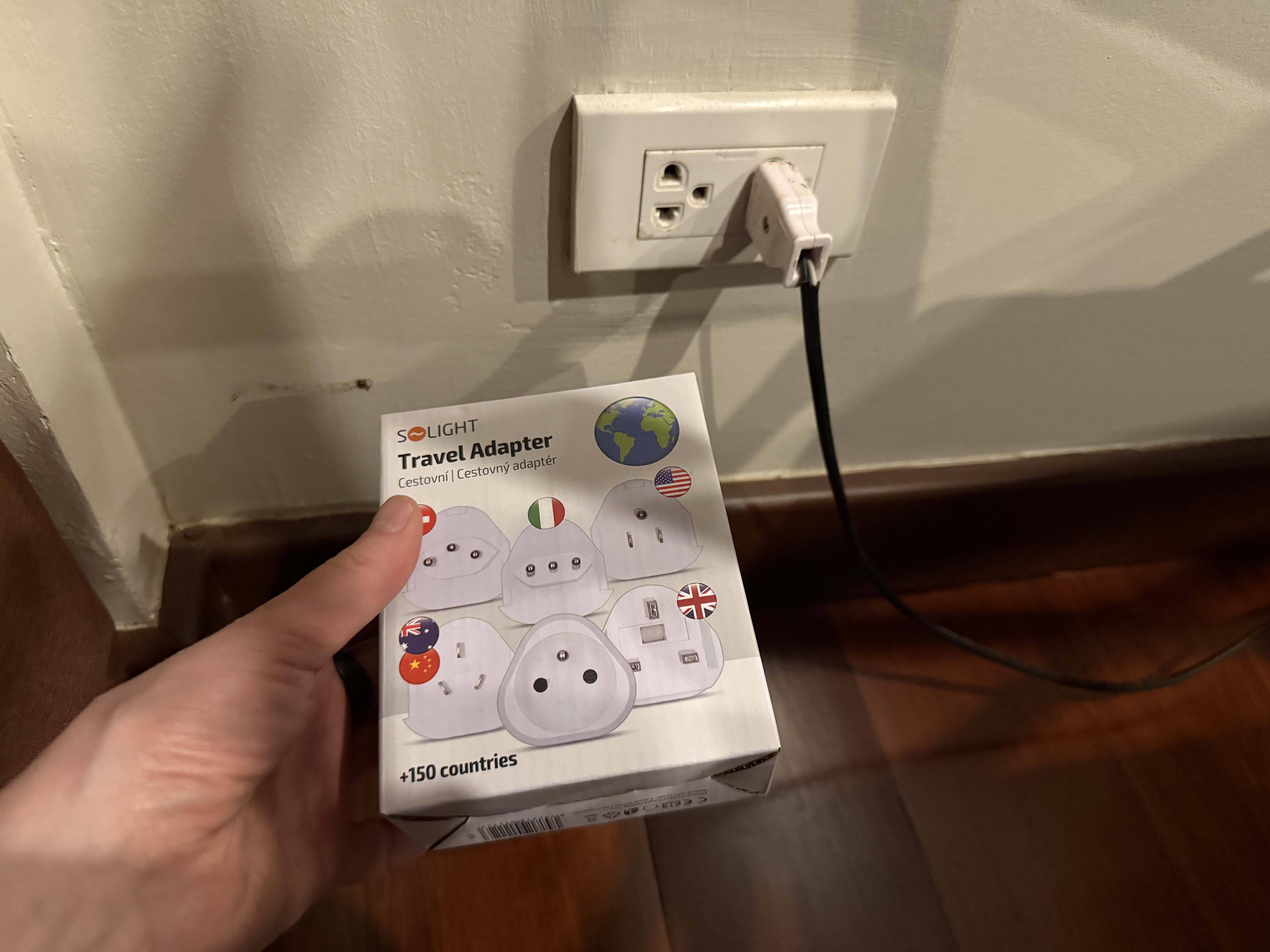 Ruka drží krabičku SOLIGHT Travel Adapter +150 countries vedle zásuvky s nabíječkou