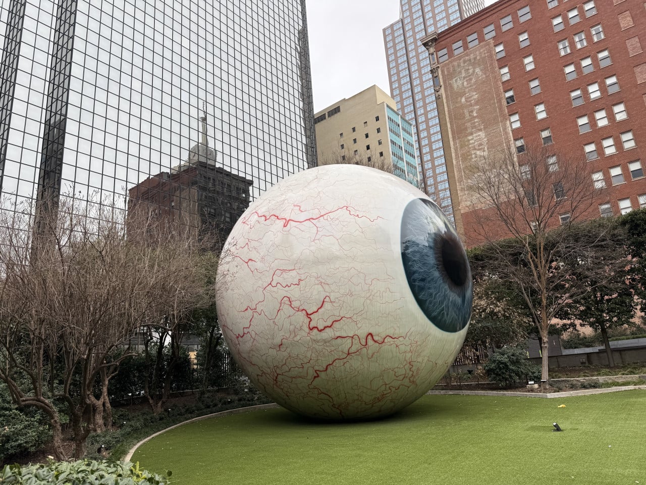 Obří socha oka na travnatém náměstí, kolem mrakodrapy a stromy