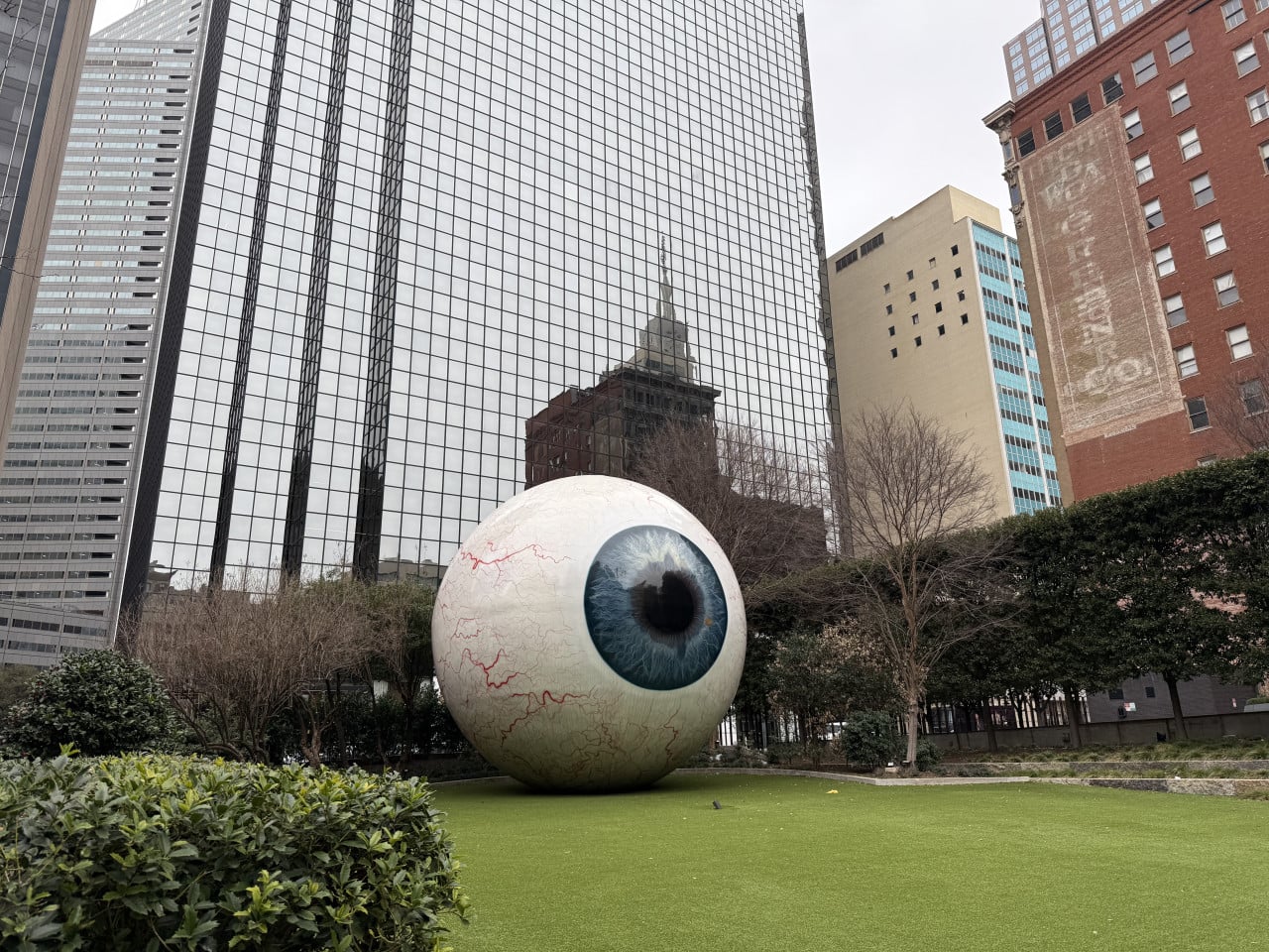 Obří socha oční bulvy na trávníku, v pozadí zrcadlové mrakodrapy downtown Dallas.