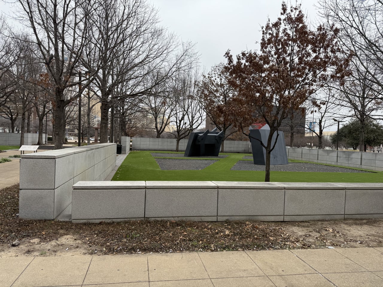Park s holými stromy, betonovými zídkami a tmavými kovovými sochami na trávníku