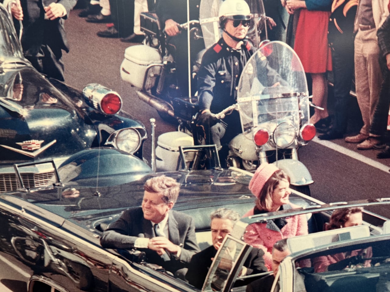 Otevřený kabriolet s politiky, policejní motorka a dav přihlížejících na ulici.