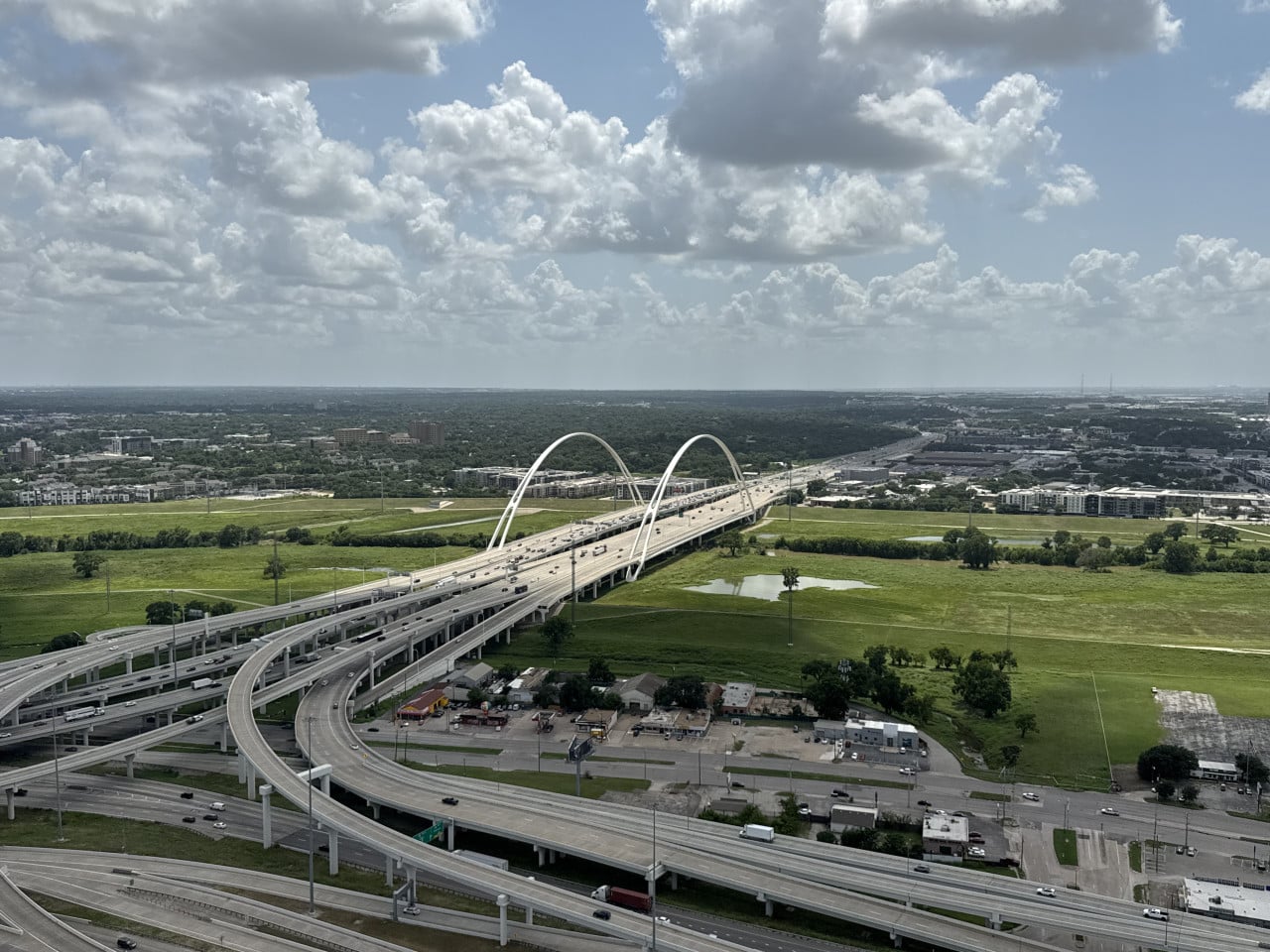 Letecký pohled na dálniční most se dvěma bílými oblouky, zelené pláně, mraky