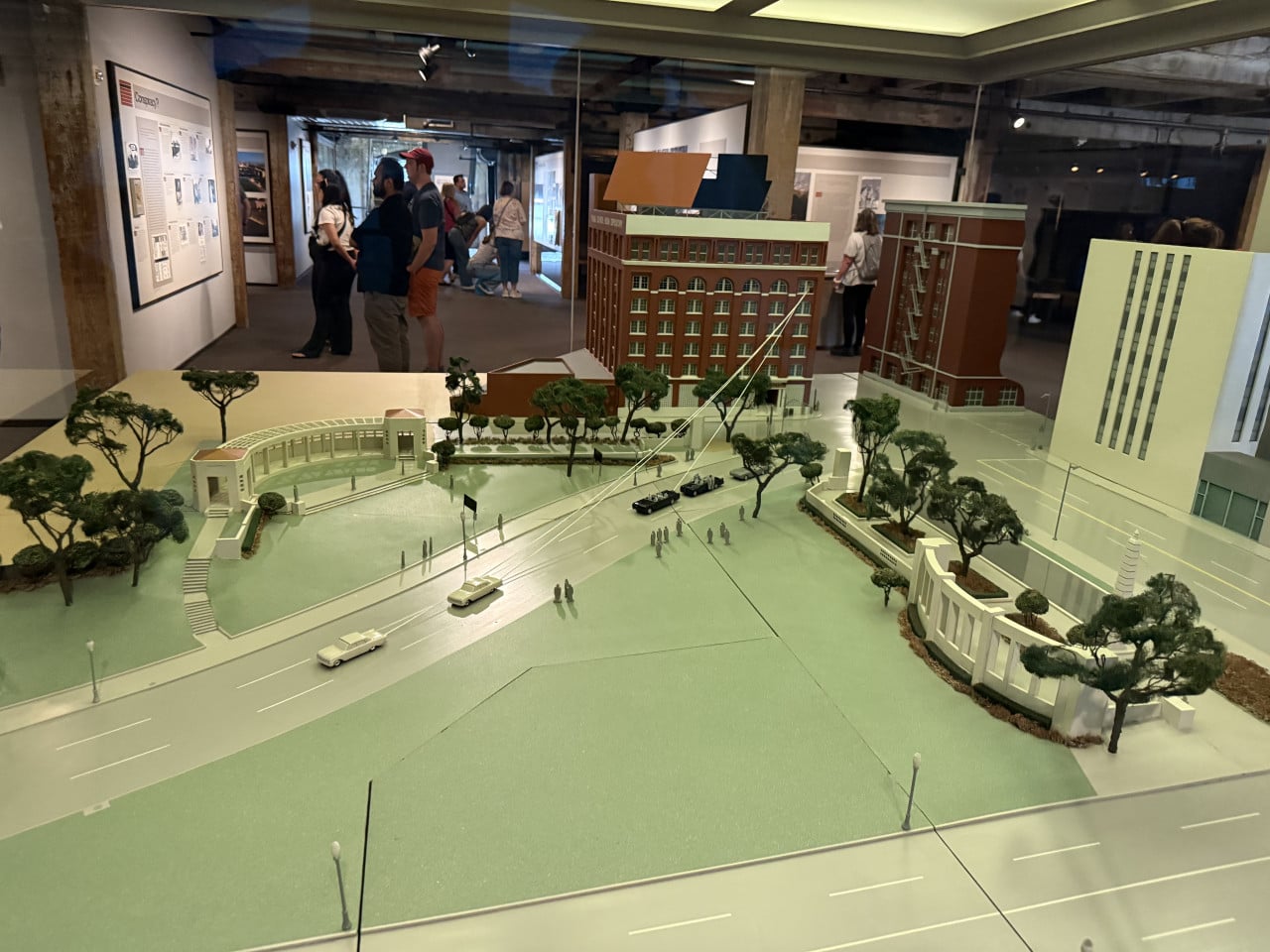 Muzejní model Dealey Plaza: silnice, park s stromy, budovy a návštěvníci v pozadí