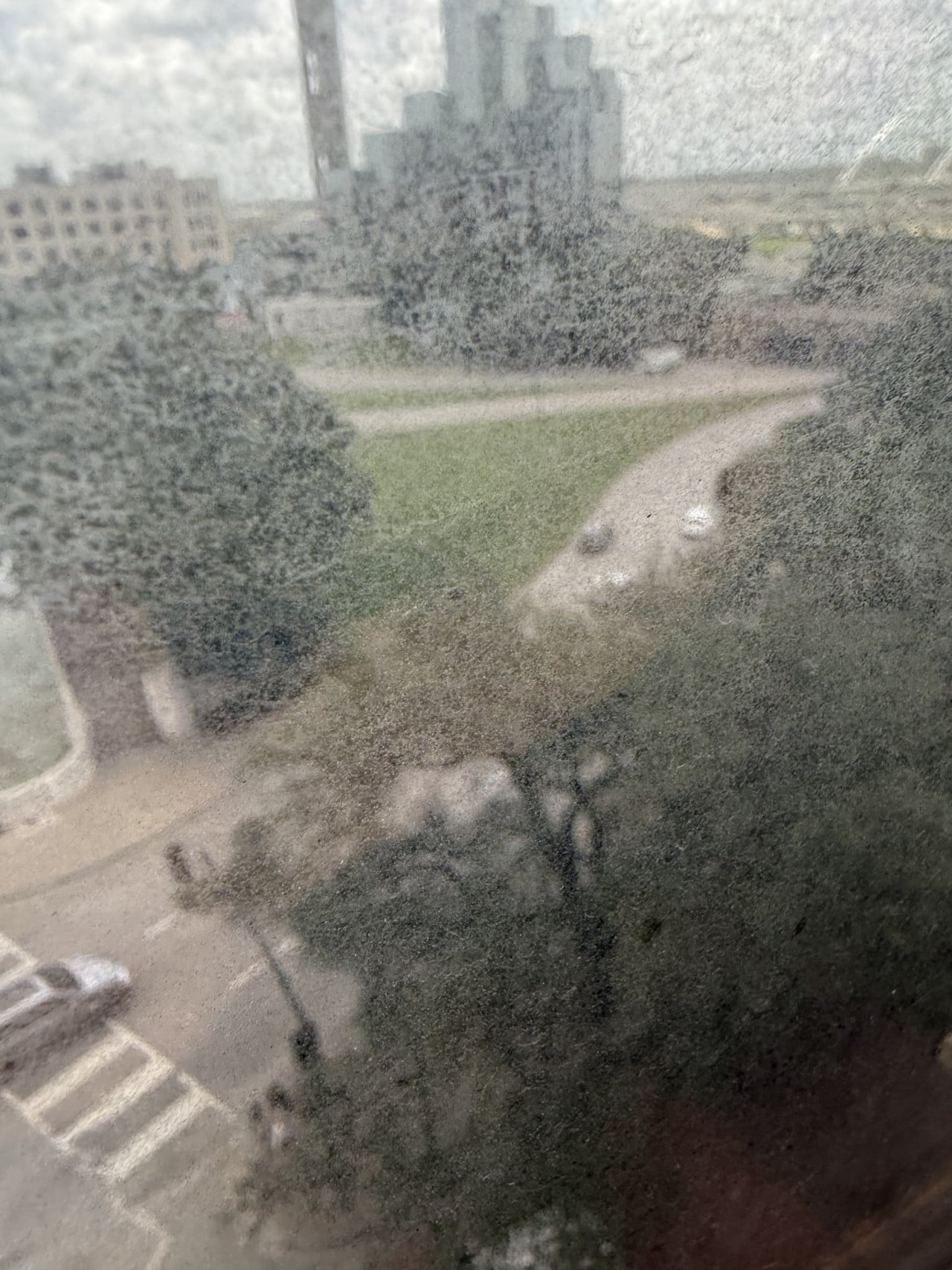 Pohled přes špinavé okno na park, cestičku, stromy a mrakodrapy v dálce