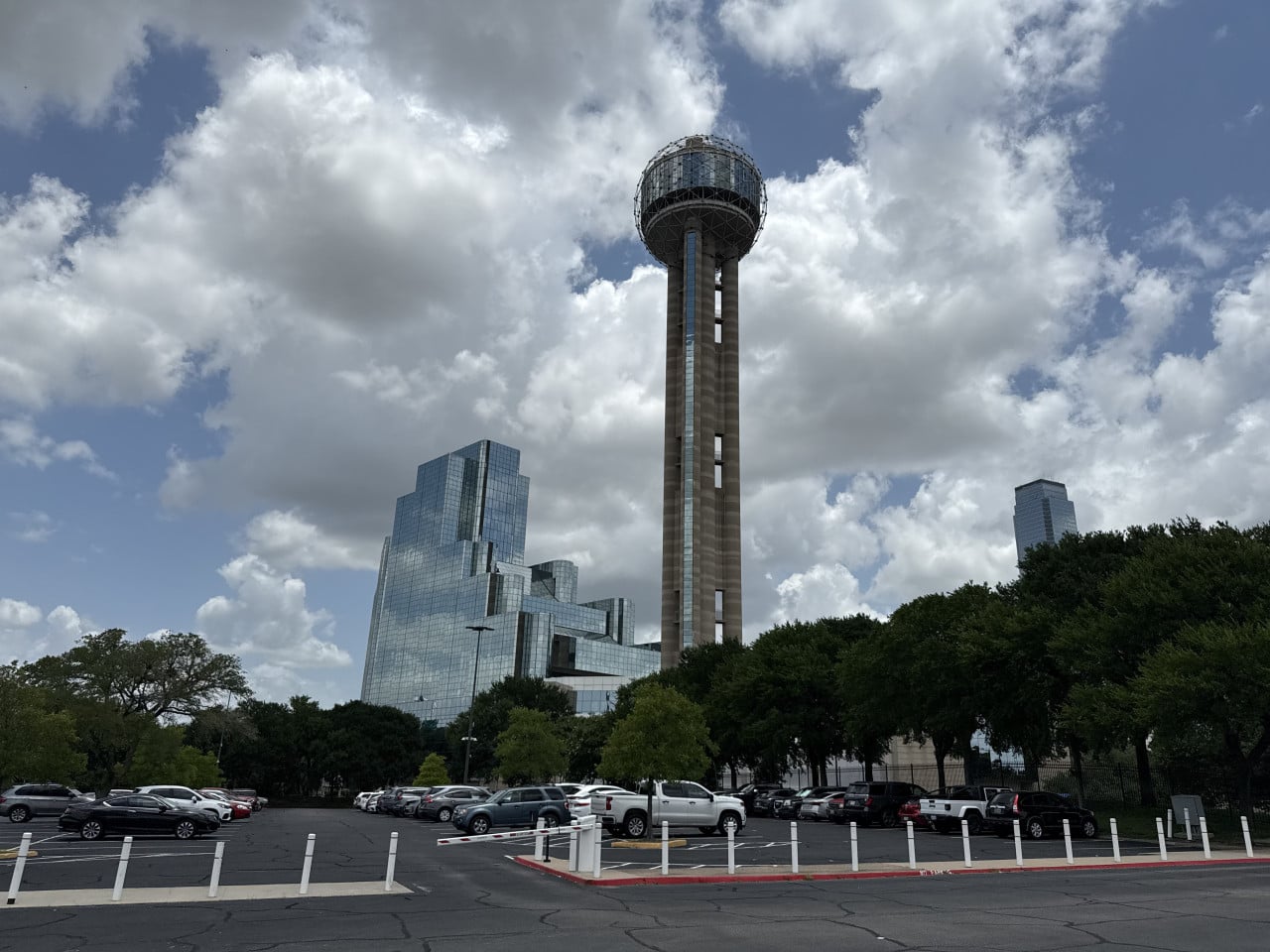 Reunion Tower a moderní prosklené mrakodrapy nad parkovištěm pod oblačnou oblohou.