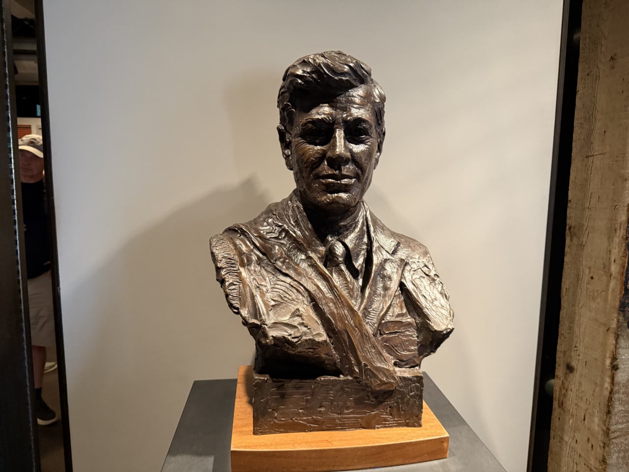 Bronzová busta muže v obleku a kravatě na podstavci v interiéru.