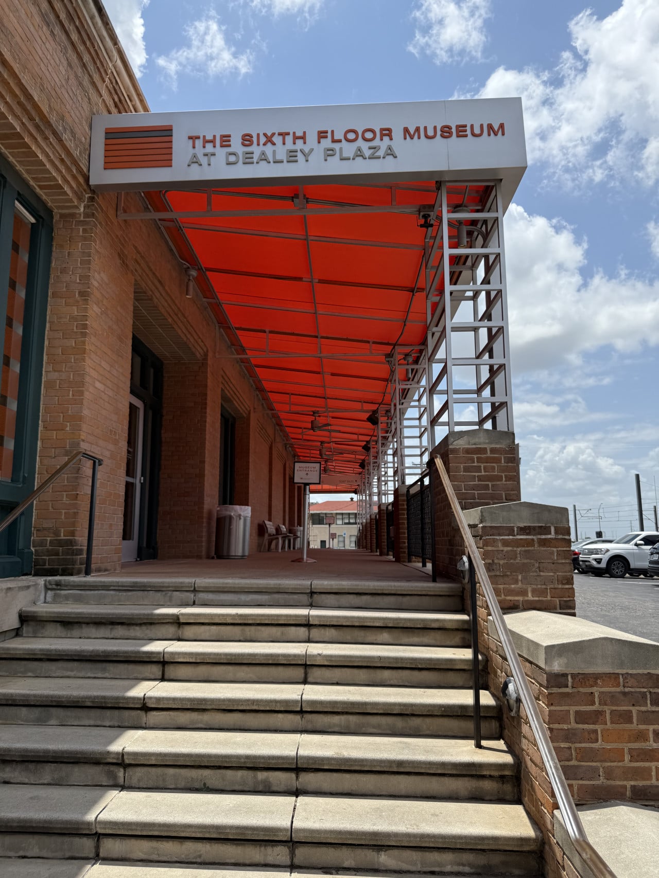Schody k cihlové budově, červený přístřešek a nápis THE SIXTH FLOOR MUSEUM AT DEALEY PLAZA.