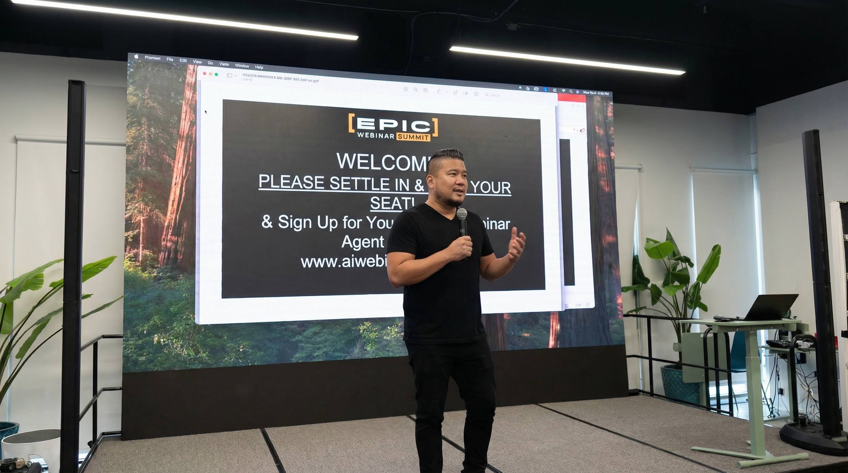 Speaker s mikrofonem před obrazovkou EPIC WEBINAR SUMMIT, WELCOME PLEASE SETTLE IN.