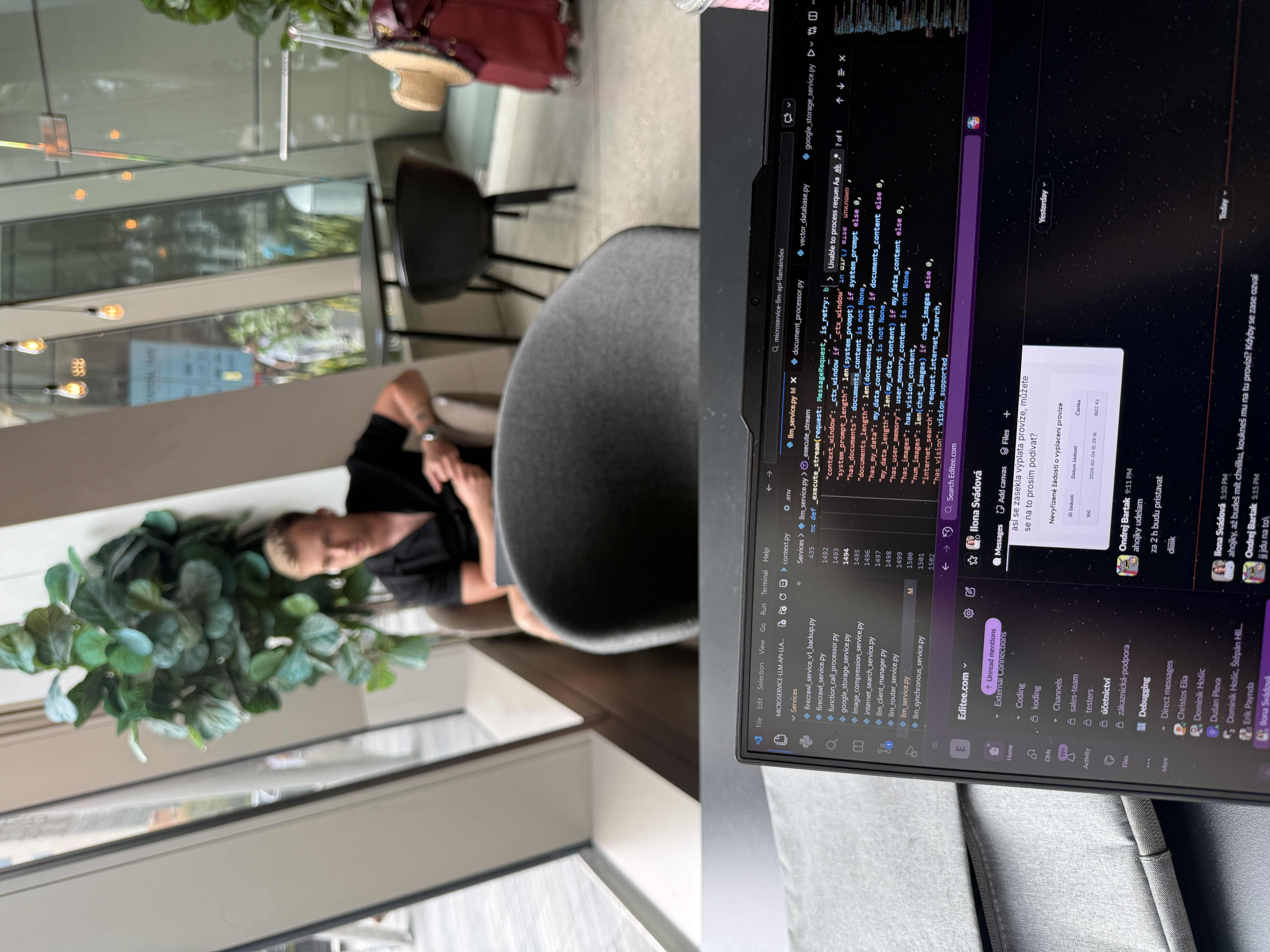 Laptop s kódem a Slackem Editee.com, vzadu rozmazaný muž v křesle.