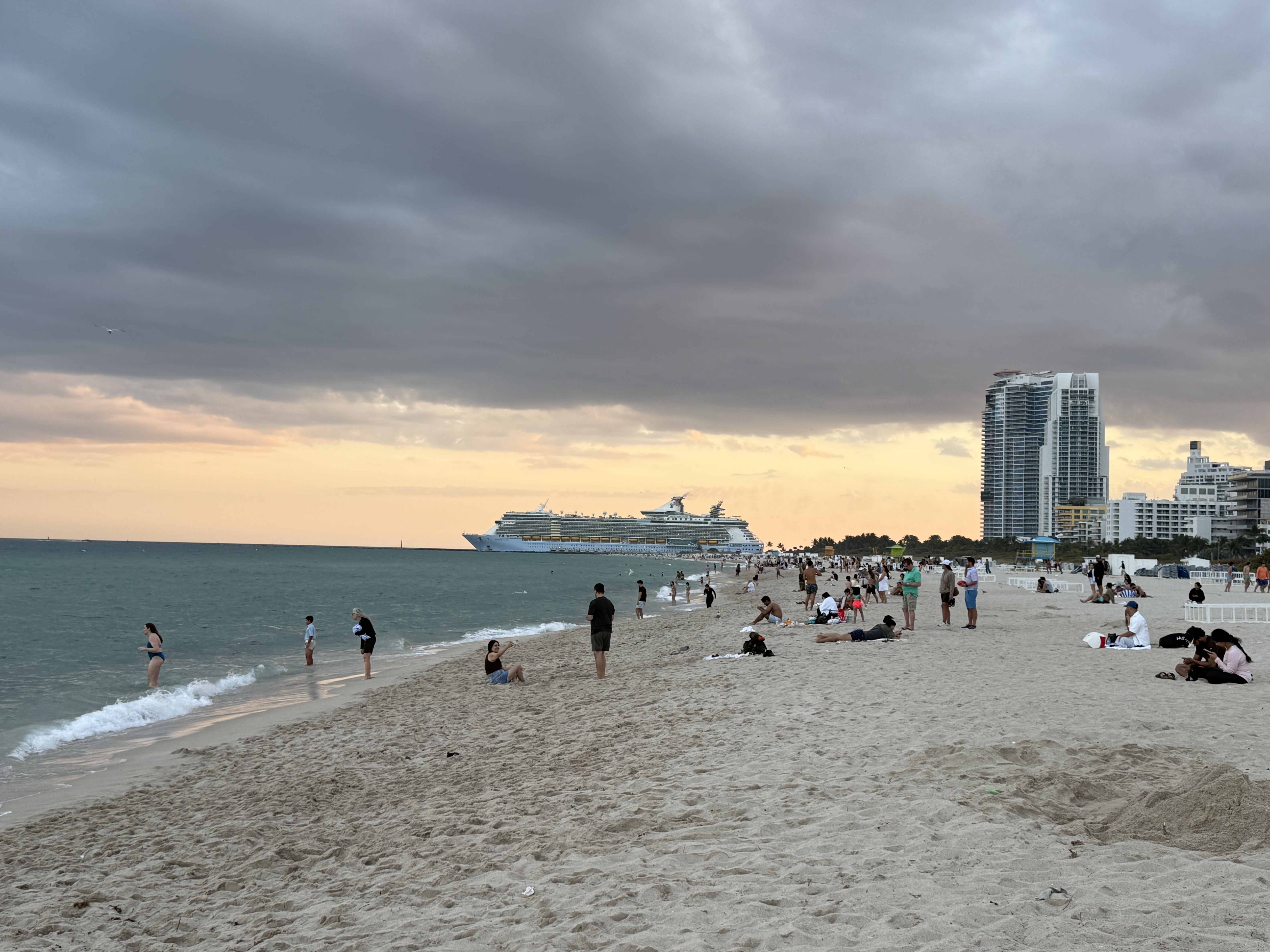 Pláž v Miami při západu slunce, lidé na písku, výletní loď a mrakodrapy