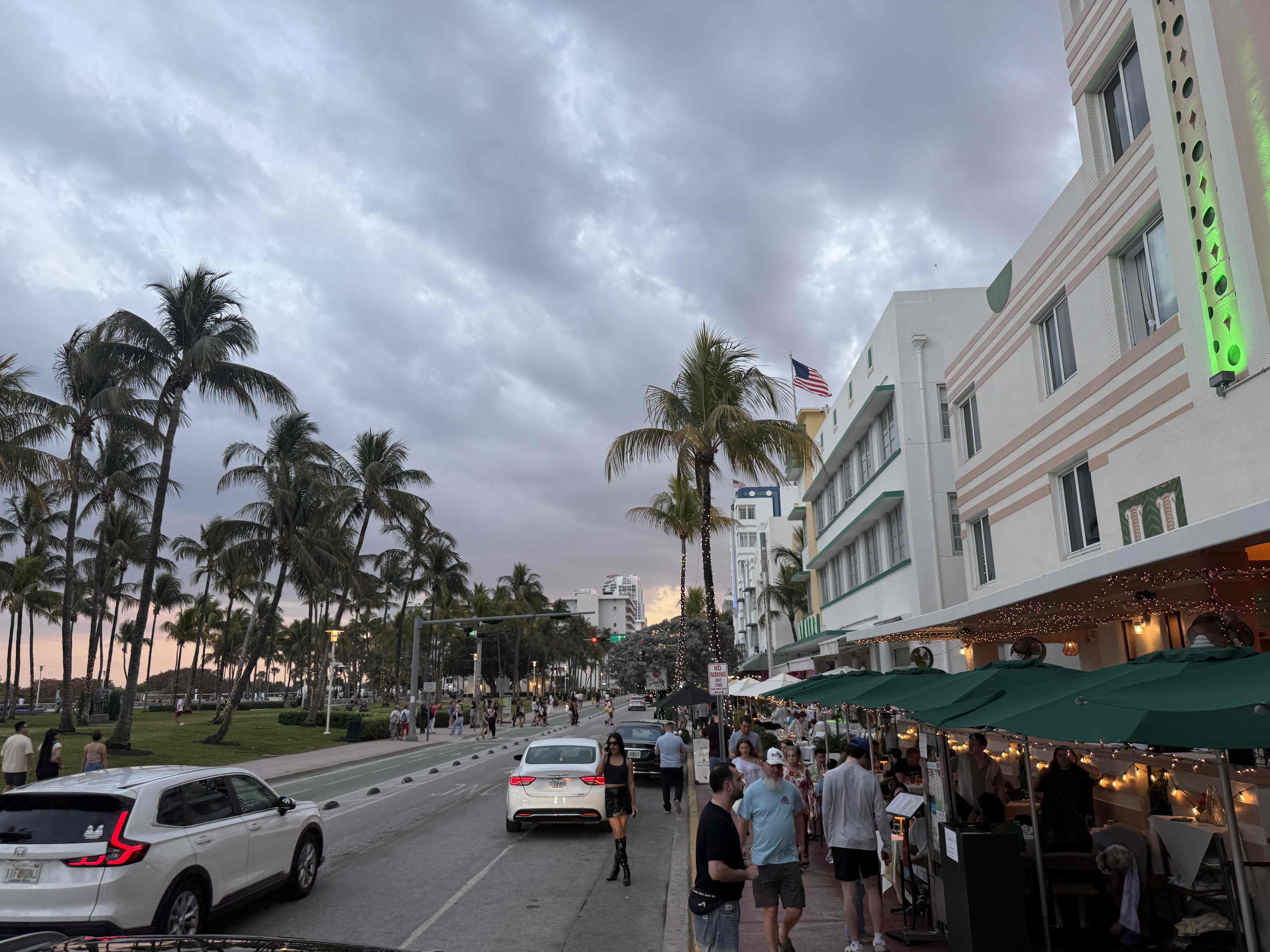 Ocean Drive v Miami: palmy, auta a lidé u restaurací pod zelenými slunečníky.