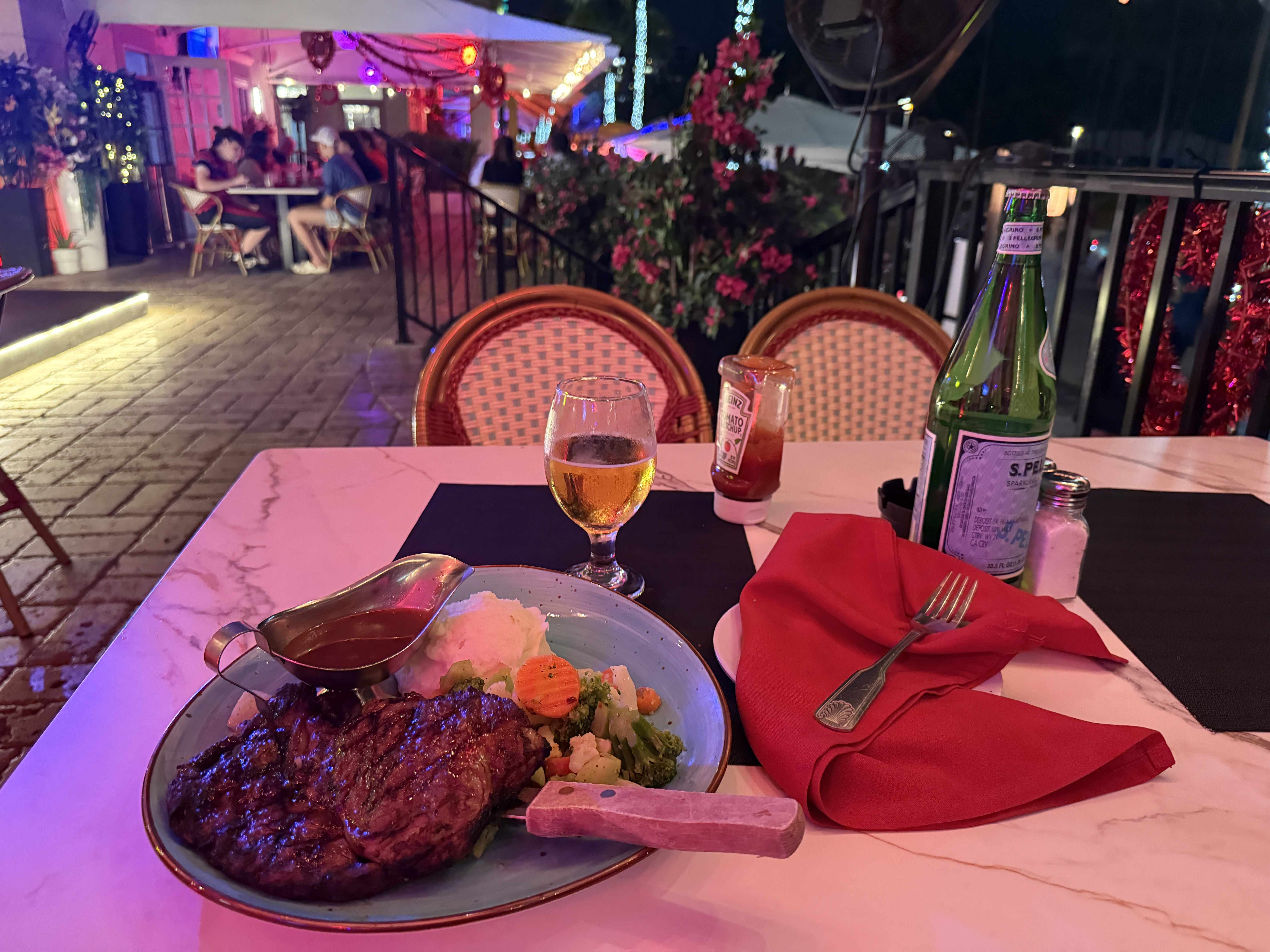 Večerní stůl v restauraci: steak, bramborová kaše, zelenina, pivo, S.Pellegrino, Heinz ketchup.