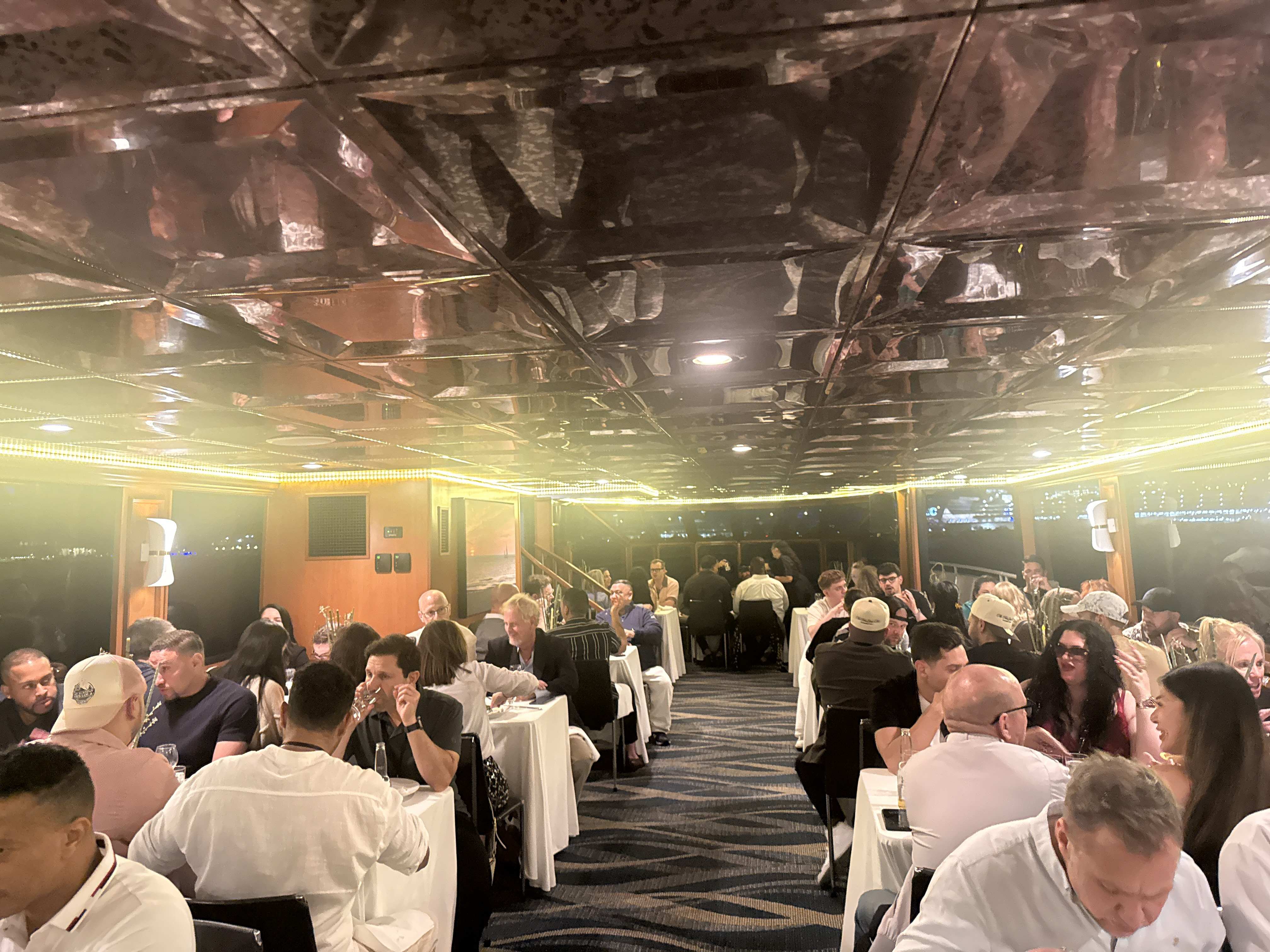 Lidé sedí u stolů v osvětleném salónu lodi, živý networking a večeře.