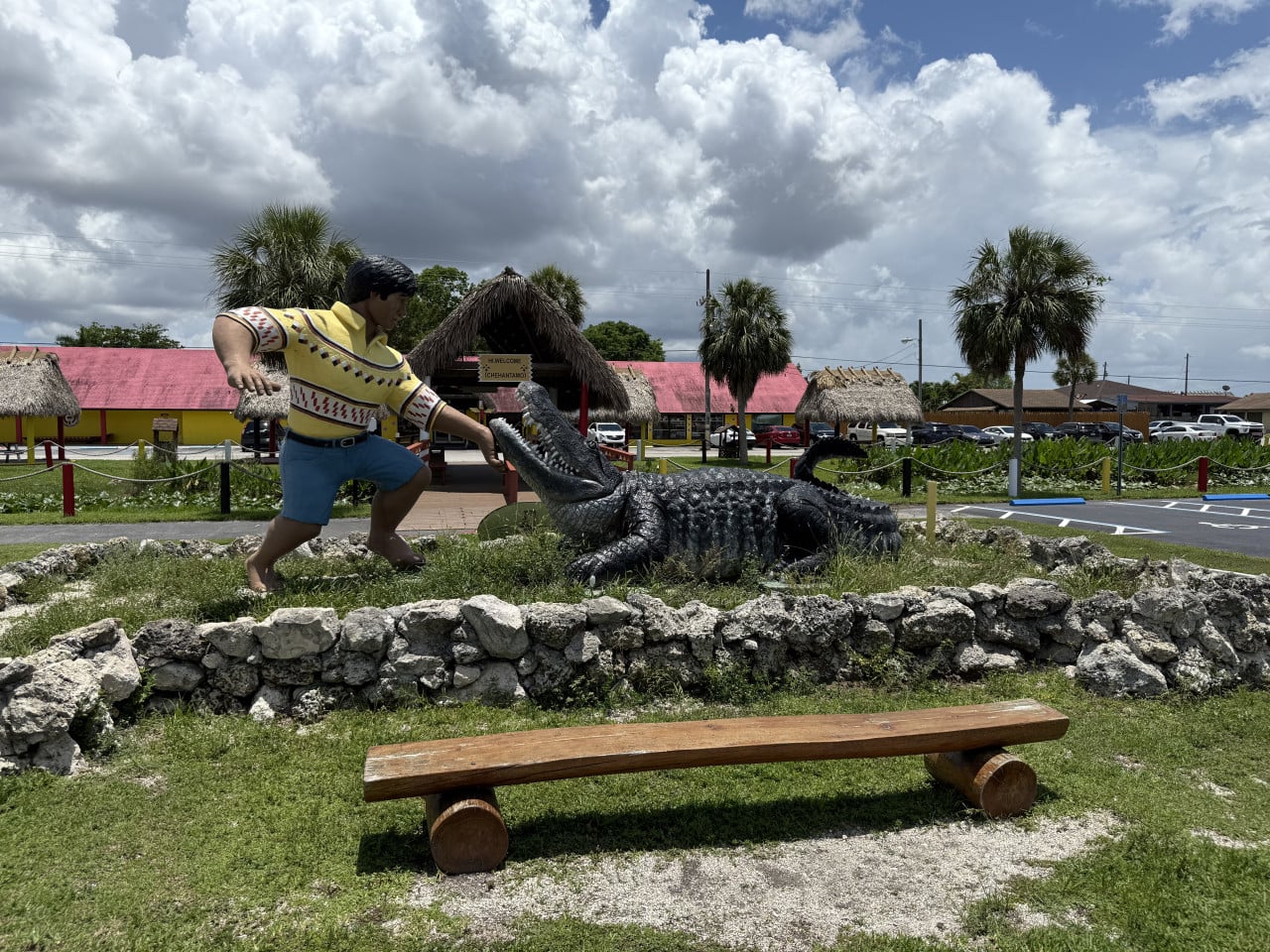 Socha muže bojujícího s aligátorem před indiánskou vesnicí a palmami.