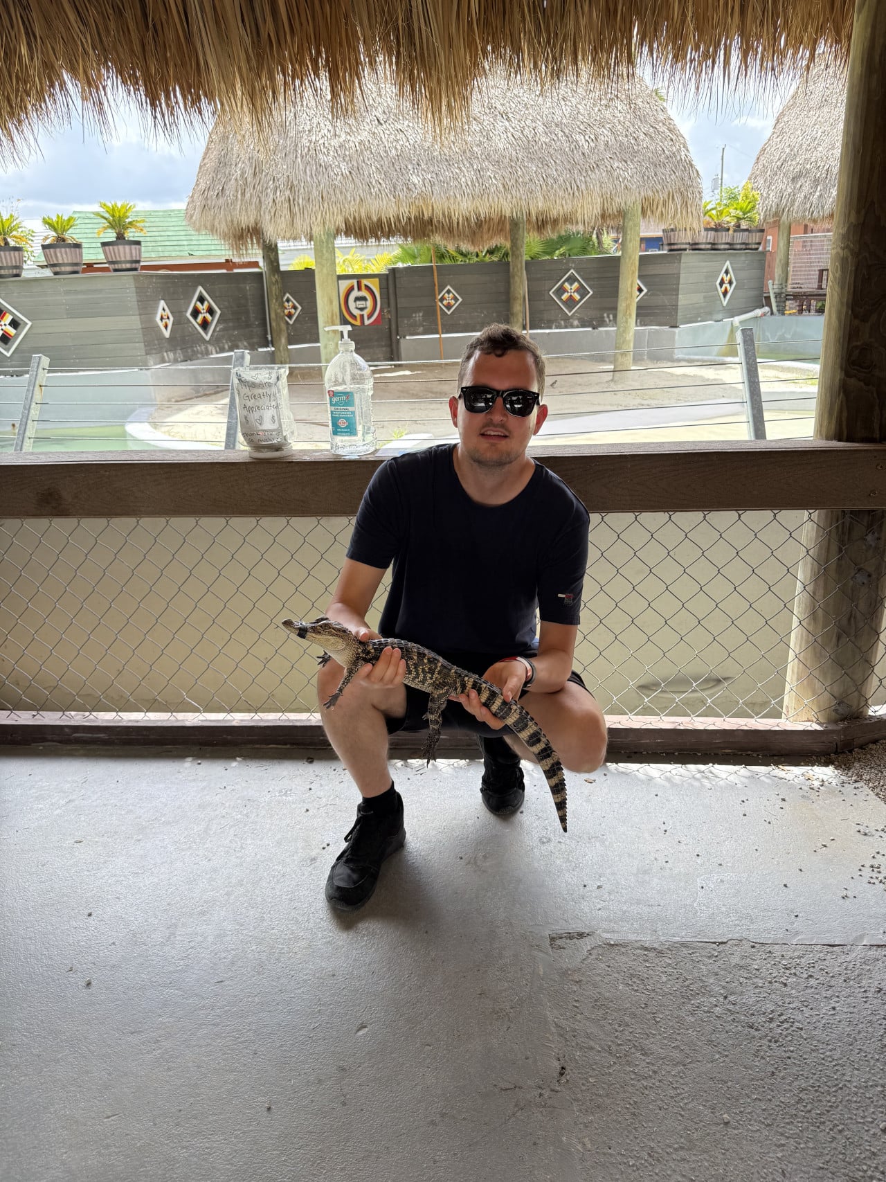 Muž v slunečních brýlích drží malého aligátora v tropickém areálu.