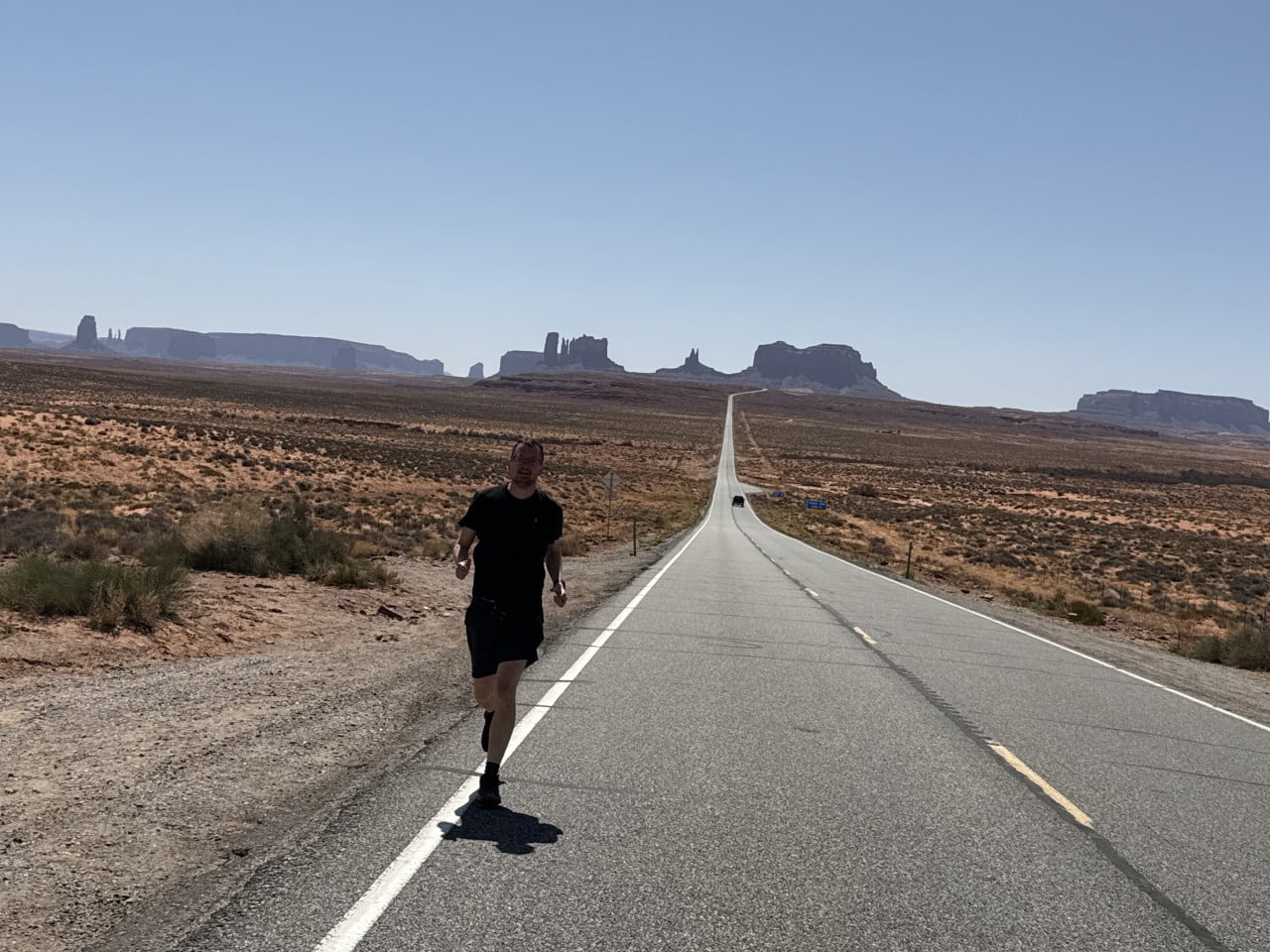 Muž běží po dlouhé rovné silnici v Monument Valley, skály v dálce.