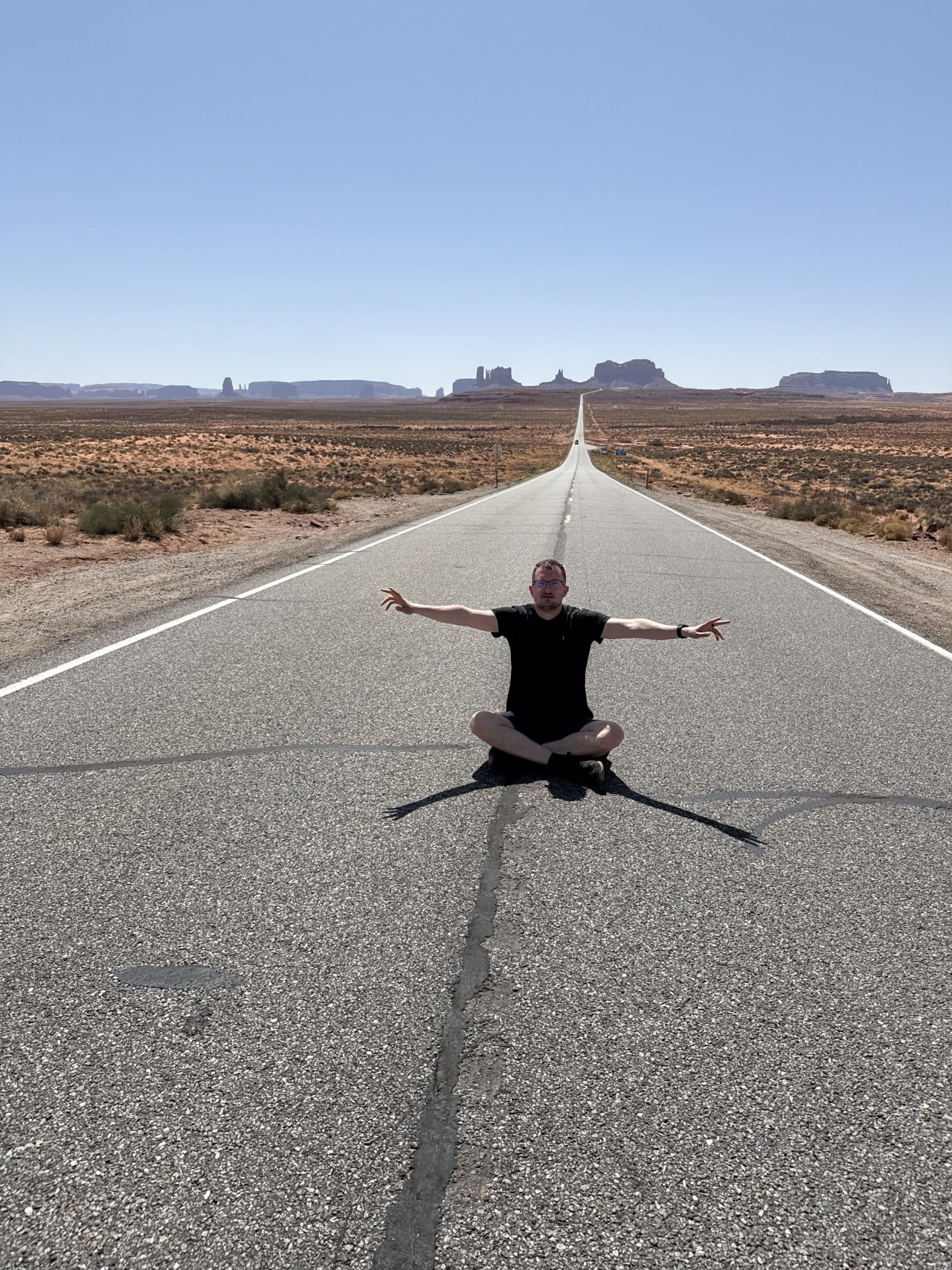 Muž sedí uprostřed rovné silnice US 163, v pozadí Monument Valley