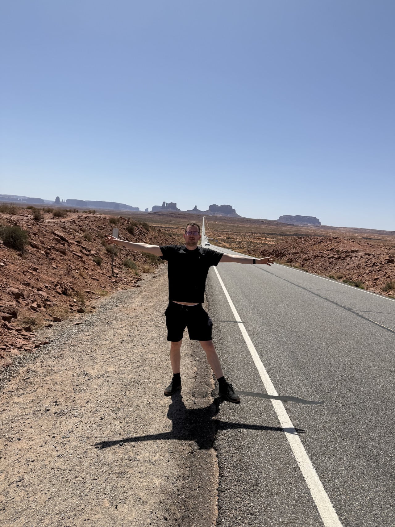 Muž s rozpaženýma rukama u rovné silnice, v dálce skály Monument Valley