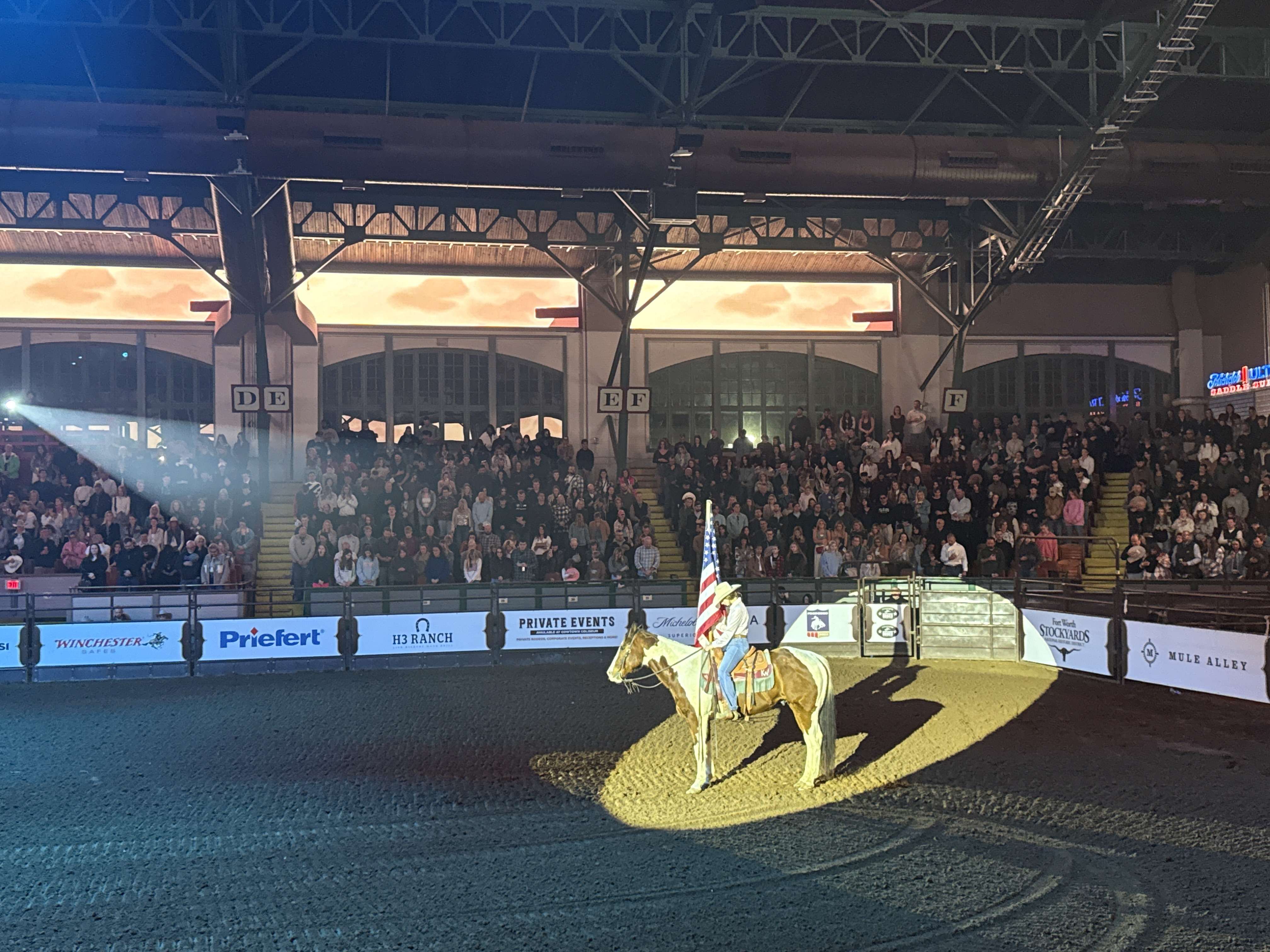 Kovboj na koni s americkou vlajkou v aréně Fort Worth Stockyards, plné tribuny.