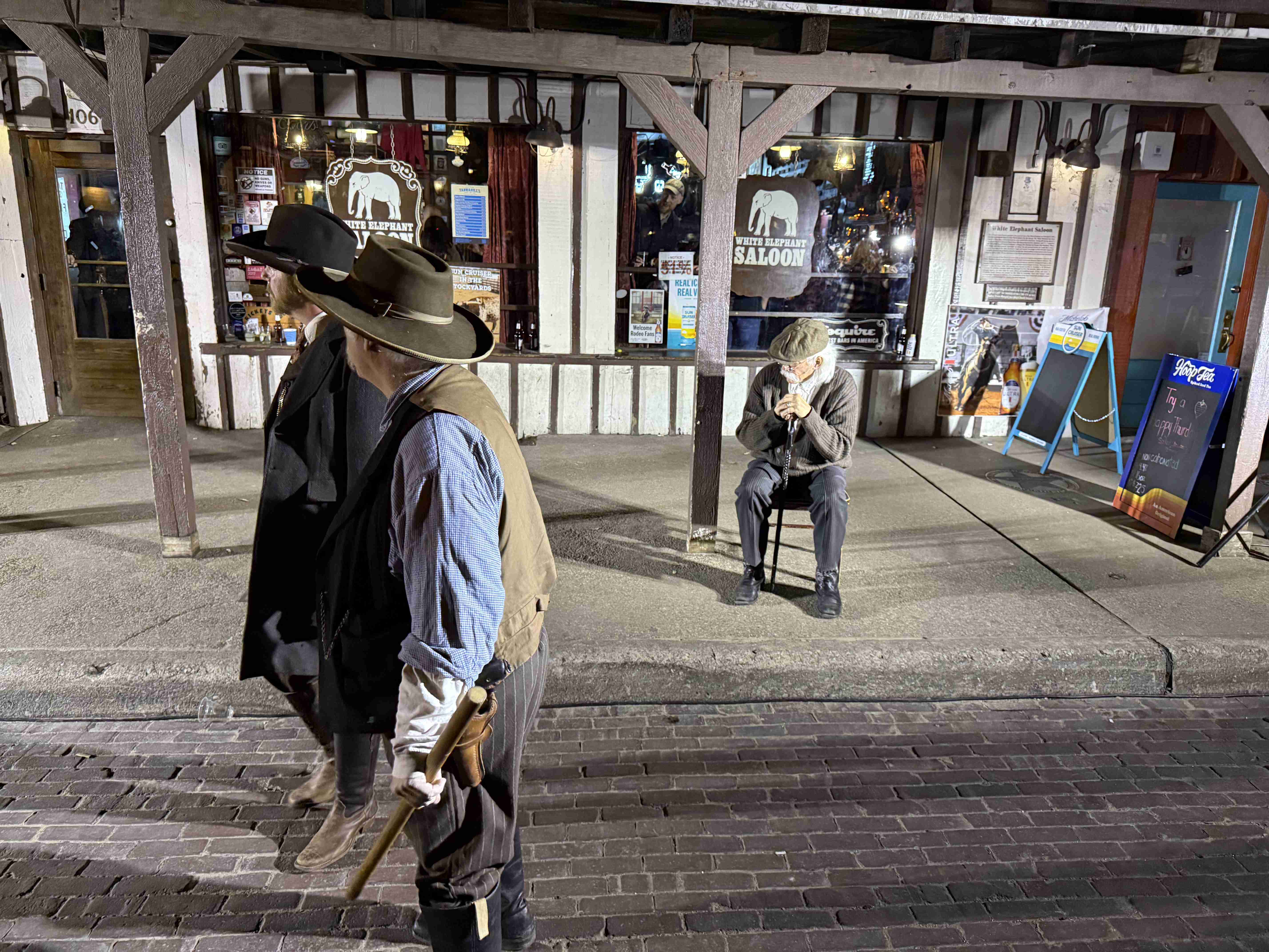 Kovbojové před White Elephant Saloon, jeden se zbraní, muž sedí opodál