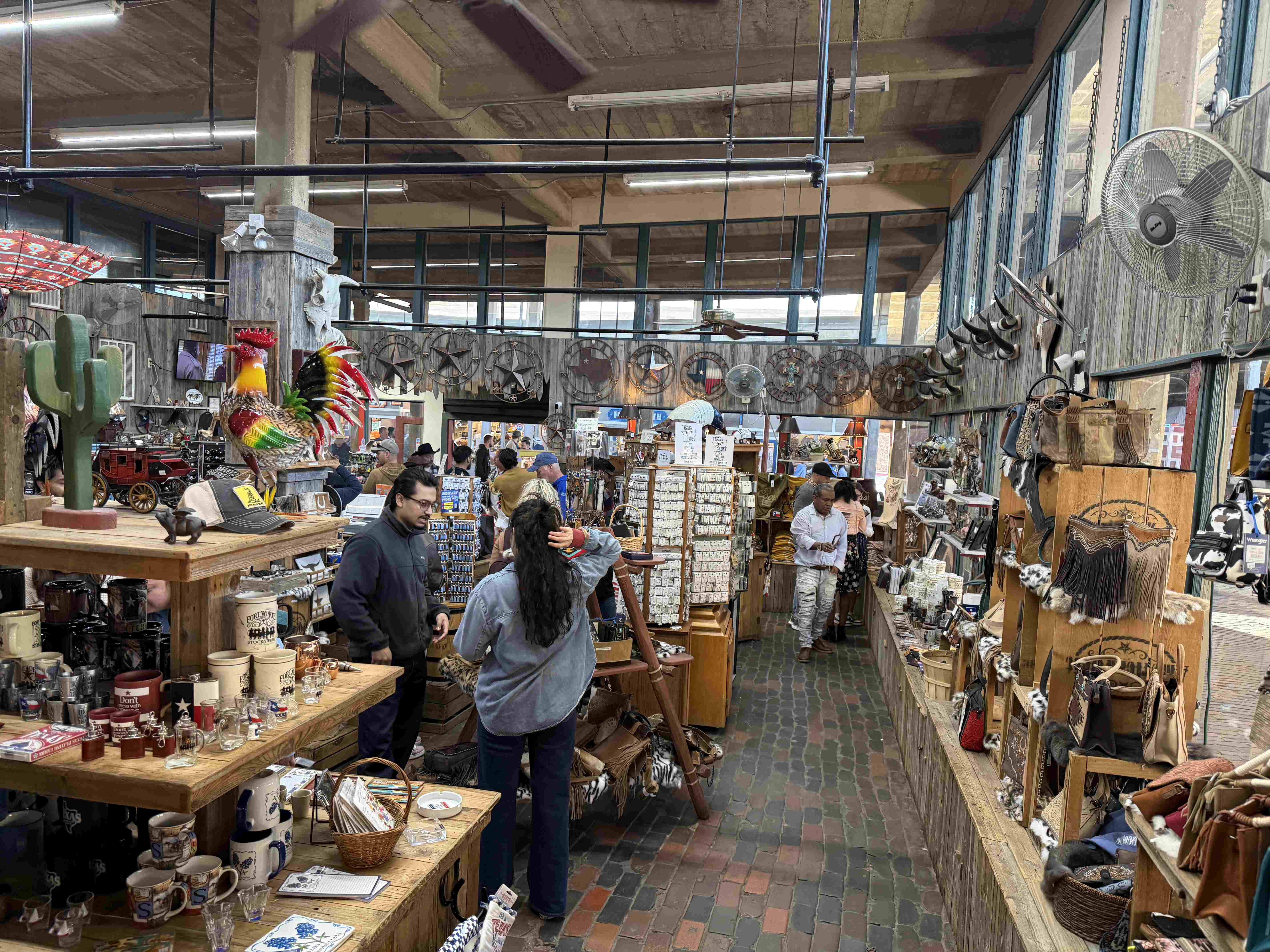 Interiér obchodu se suvenýry, lidé nakupují; hrnky s nápisem Fort Worth Stockyards.