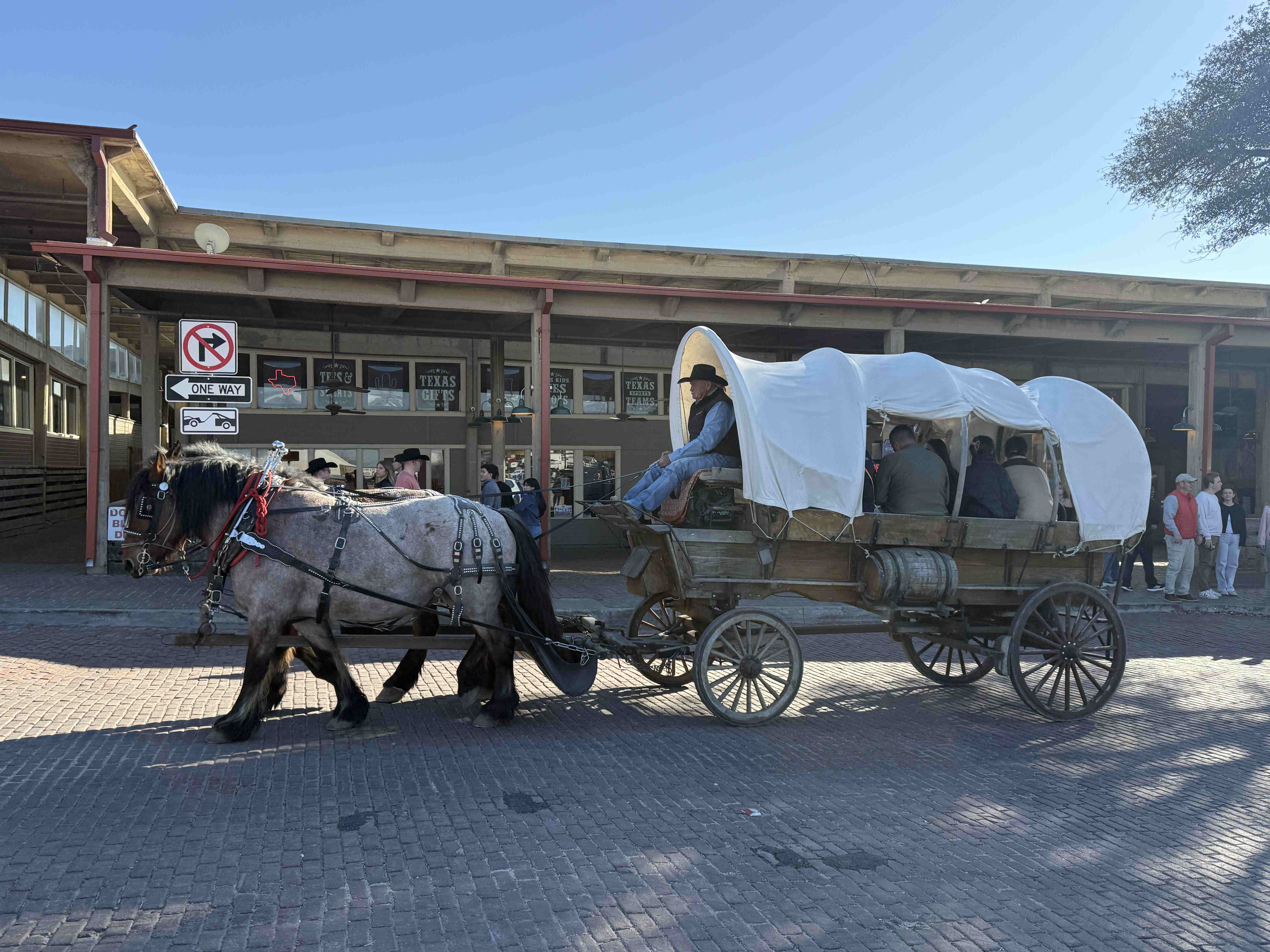 Kůň táhne krytý vůz s pasažéry; v pozadí cedule ONE WAY.