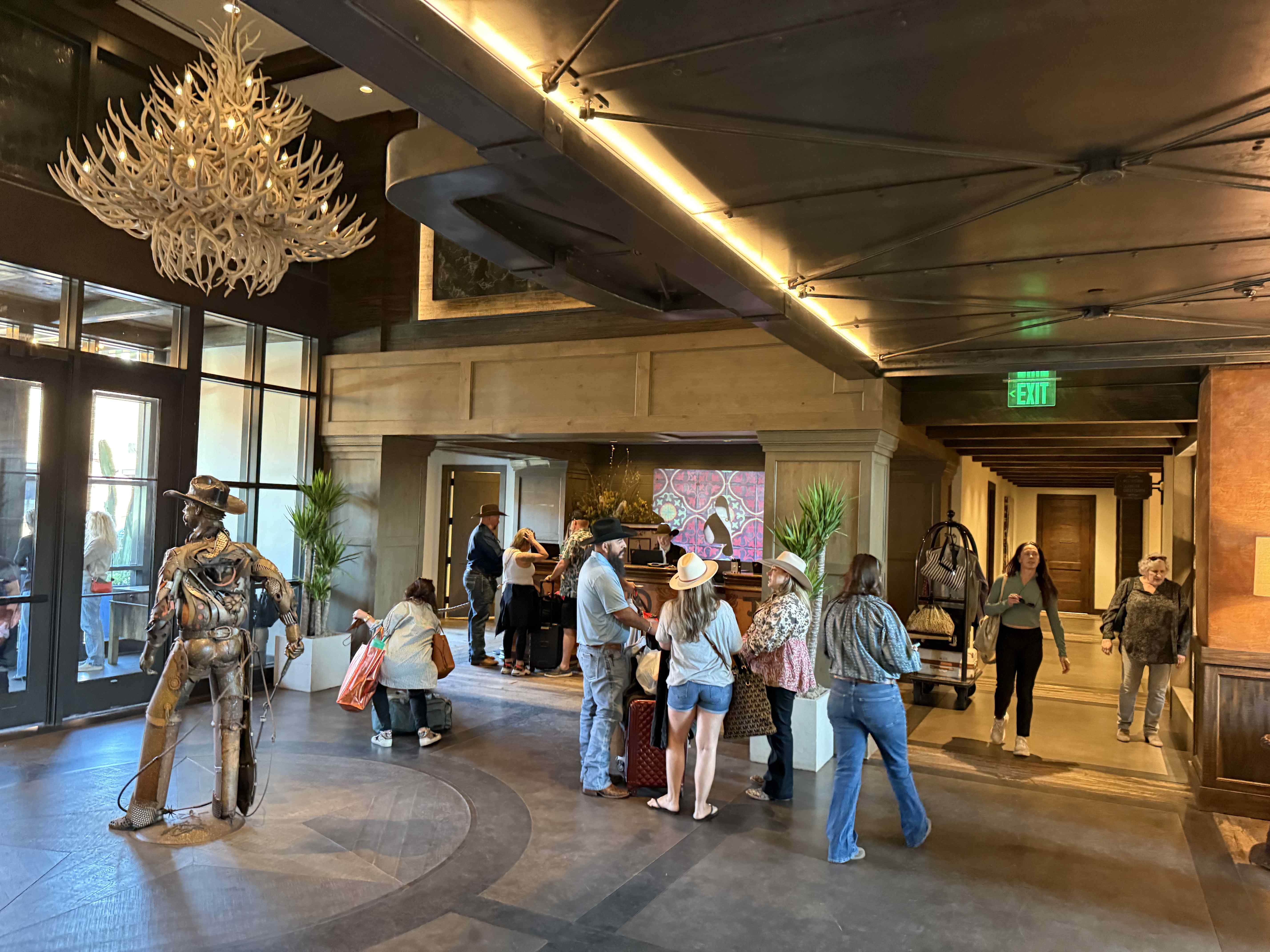 Hotelová lobby s kovbojskou sochou, lustry z paroží, lidé u recepce, EXIT