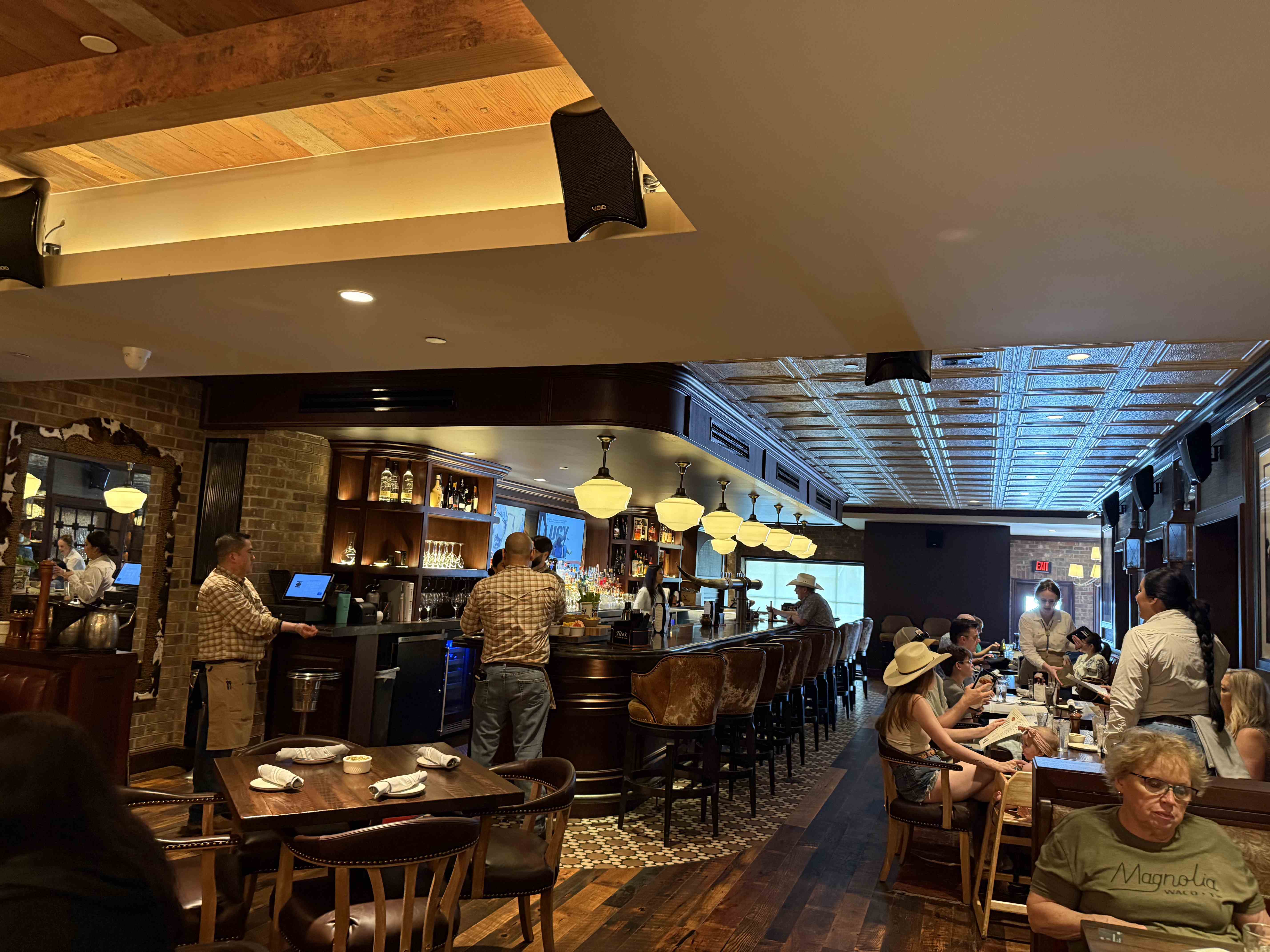 Interiér saloonu: dlouhý bar, barové stoličky, hosté v kovbojských kloboucích, nápis EXIT.