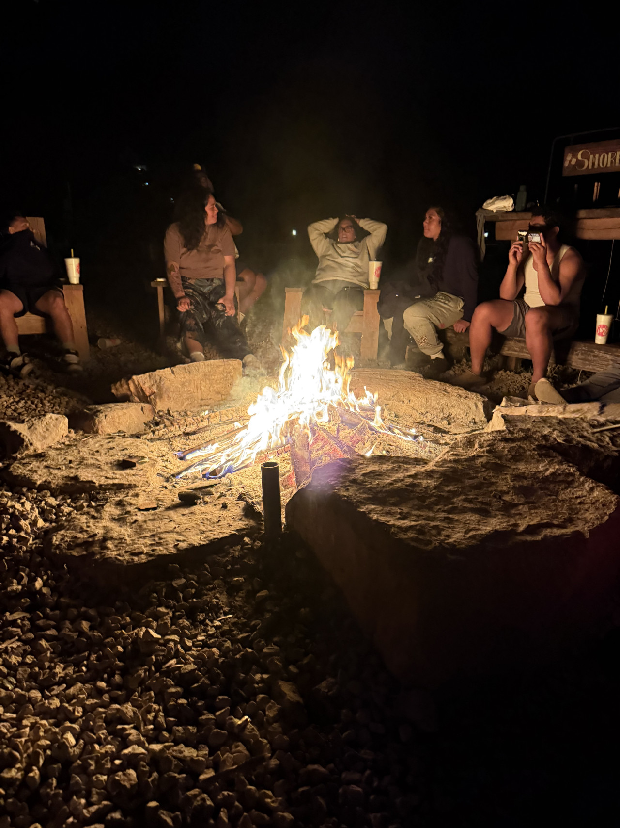 Skupina lidí sedí v noci u táboráku na dřevěných židlích; vzadu cedule S’MORE