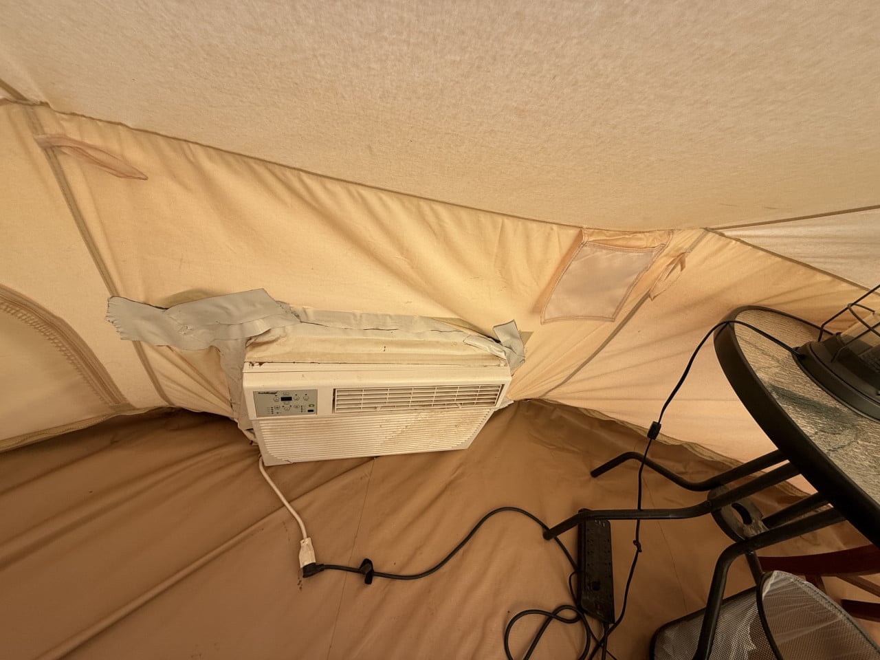Okenní klimatizace zalepená v plátně stanu, kabely na zemi a židle se stolkem
