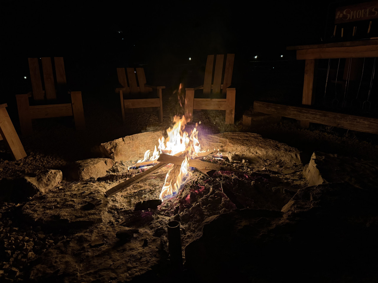 Noční táborák v kamenném ohništi, dřevěné židle kolem, cedule S’mores.