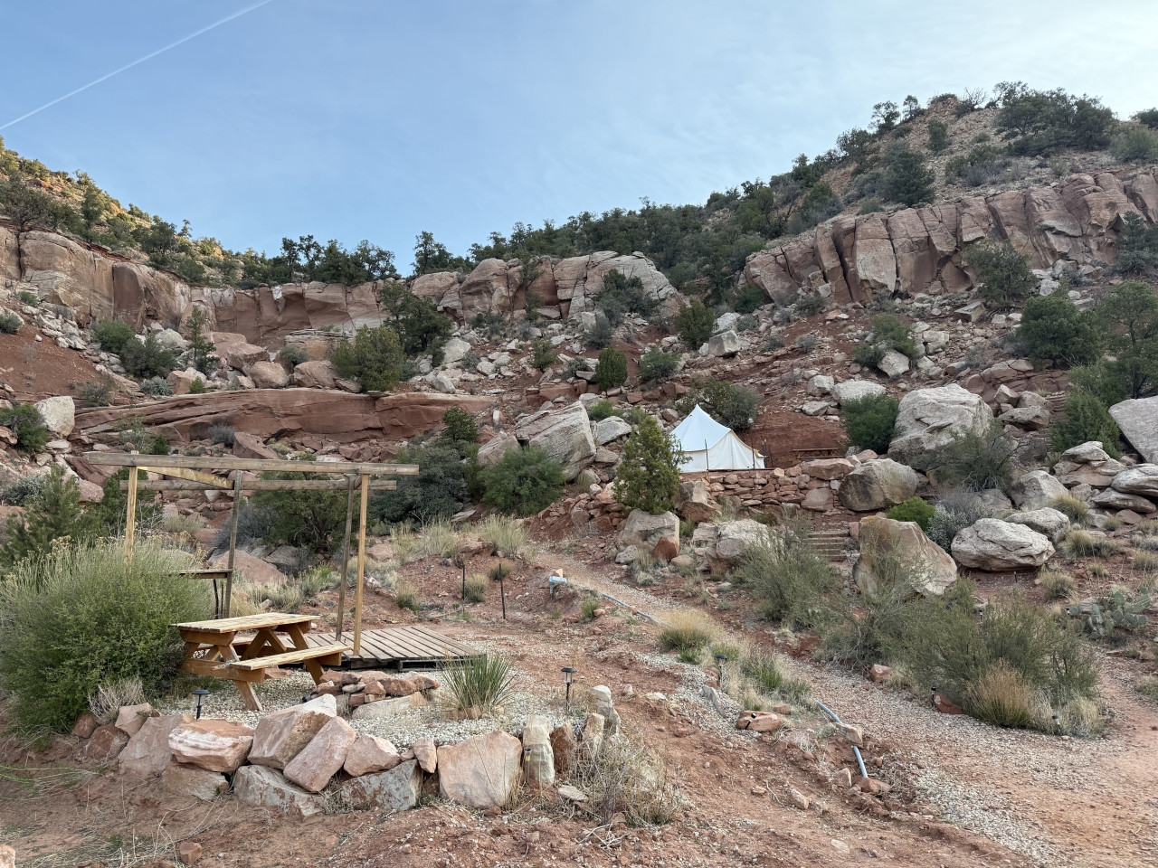 Bílý glampingový stan v kaňonu, skály, keře, pergola a piknikový stůl.