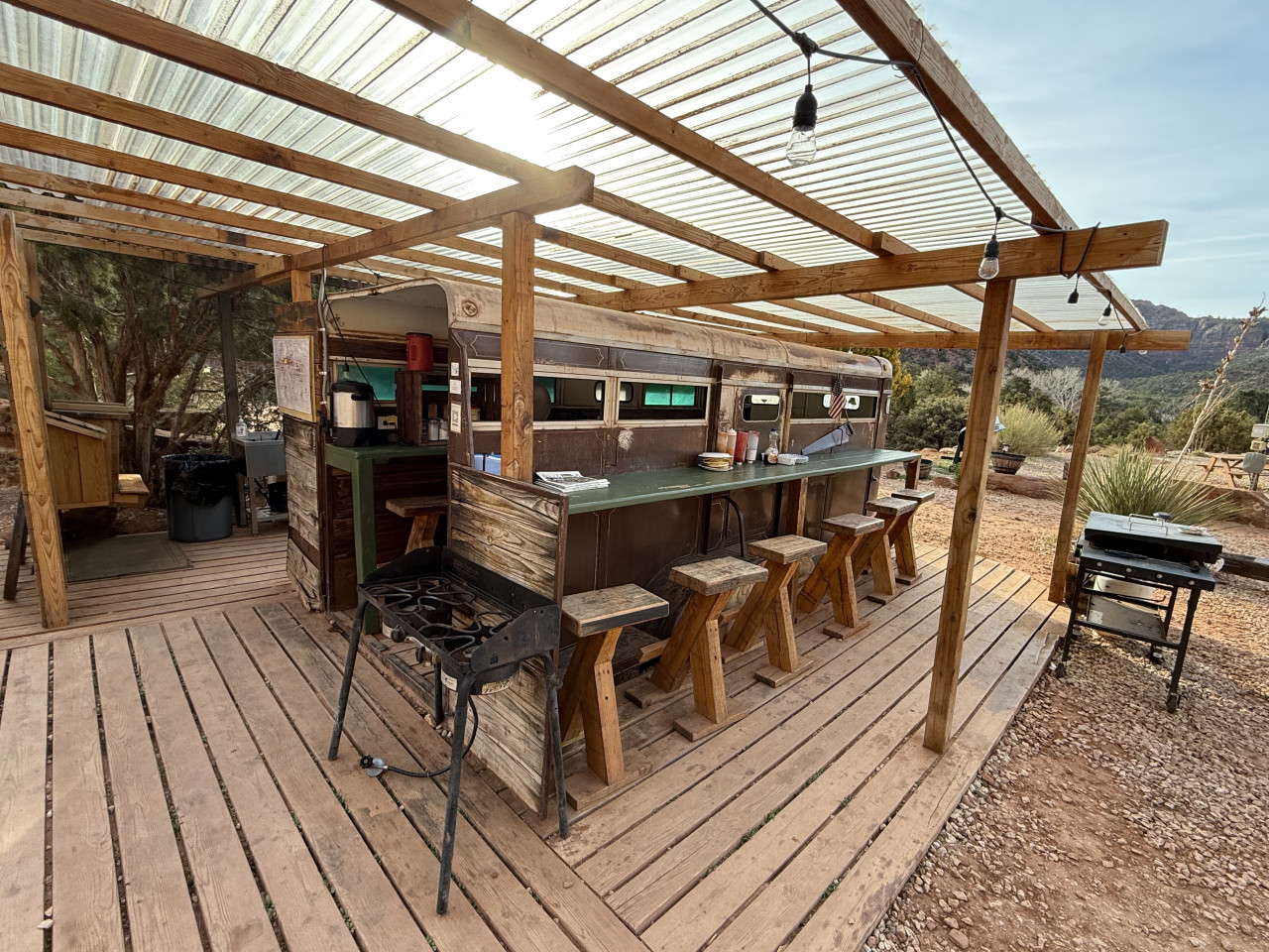 Venkovní bar z přestavěného autobusu pod pergolou, stoličky a gril.