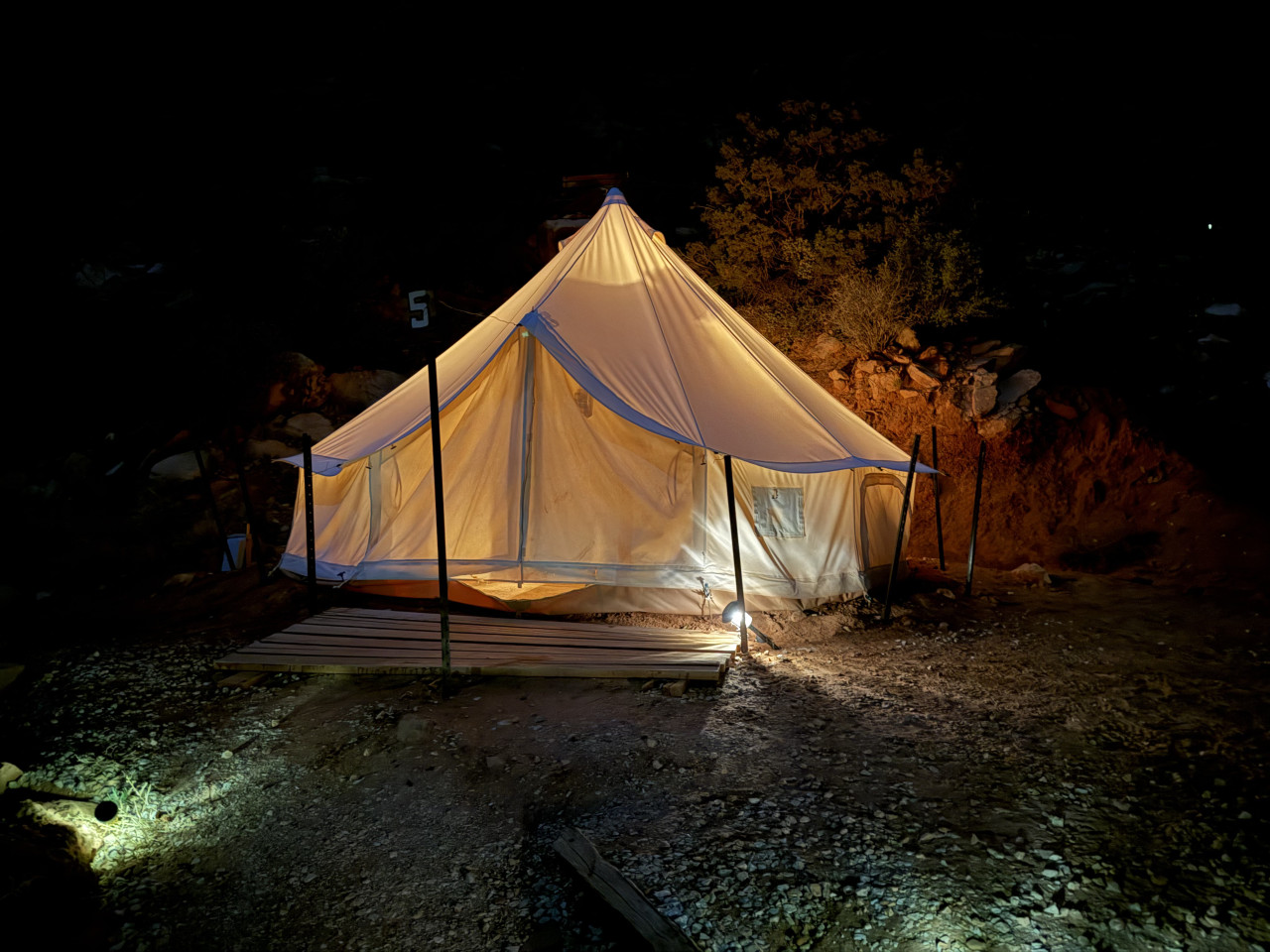 Osvětlený glampingový stan v noci u skály, cedule 5 vedle.
