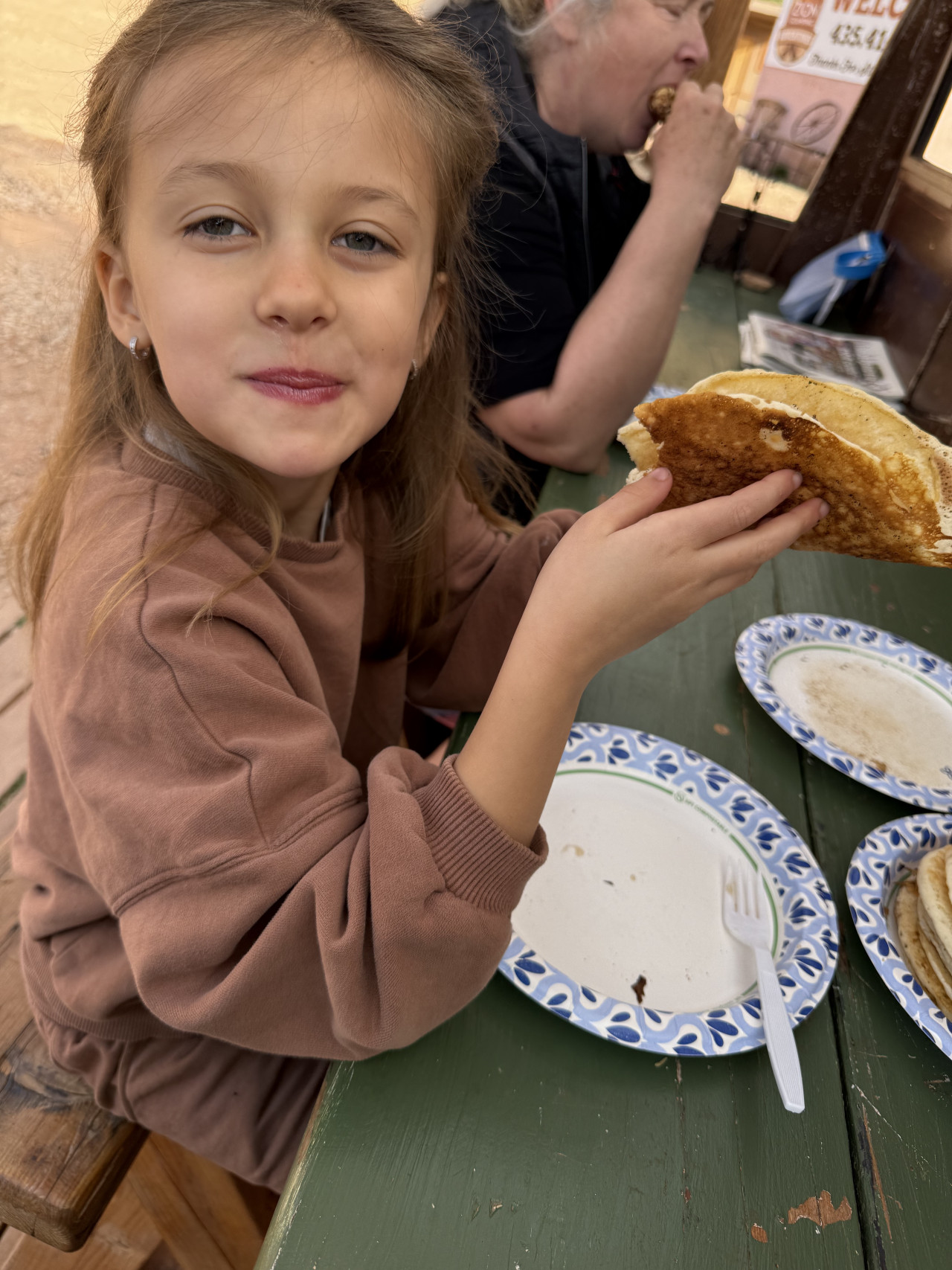 Dívka u piknikového stolu drží plněnou tortillu; v pozadí žena jí, nápis WELCOME.