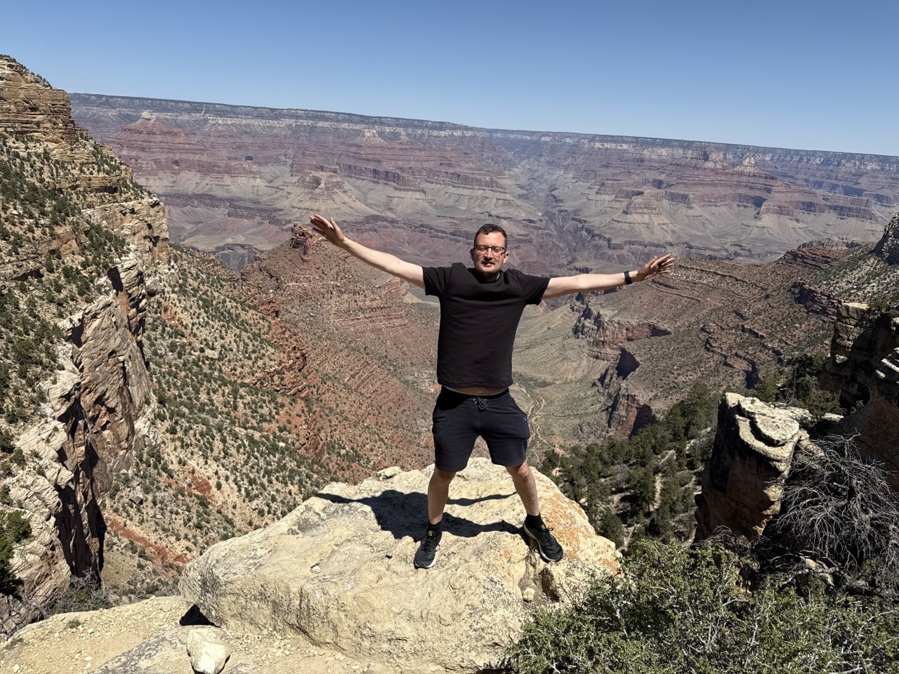 Muž stojí na skalním výběžku s rozpaženýma rukama nad Grand Canyonem.