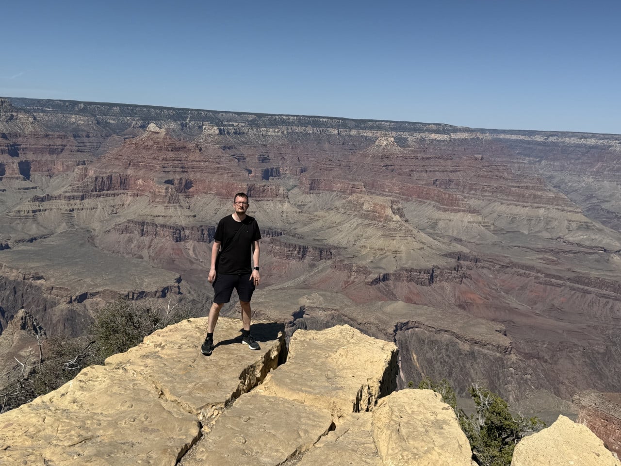 Muž stojí na skalním výběžku nad Grand Canyonem pod modrou oblohou.