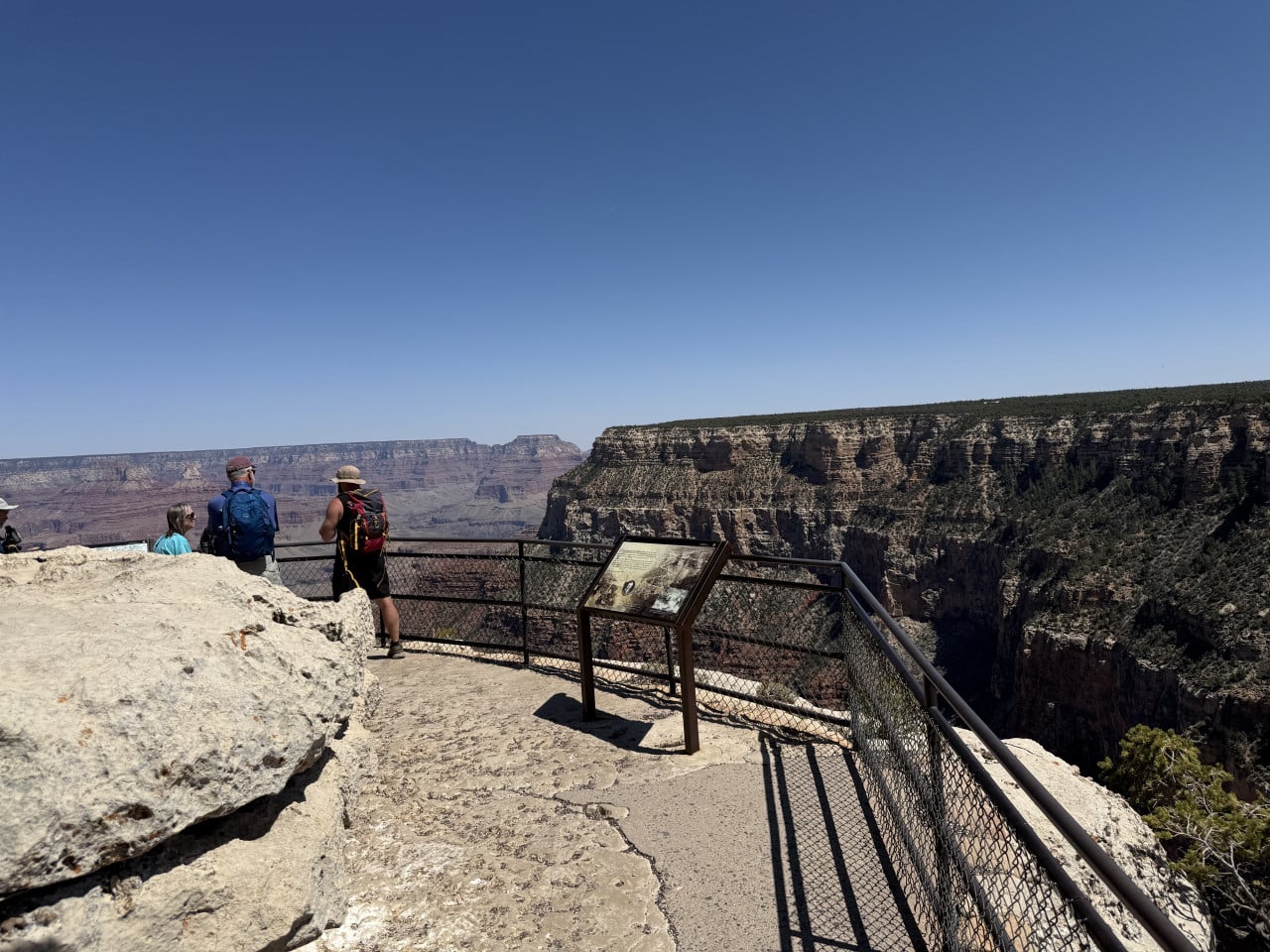 Vyhlídka na Grand Canyon s turisty u zábradlí a informační tabulí